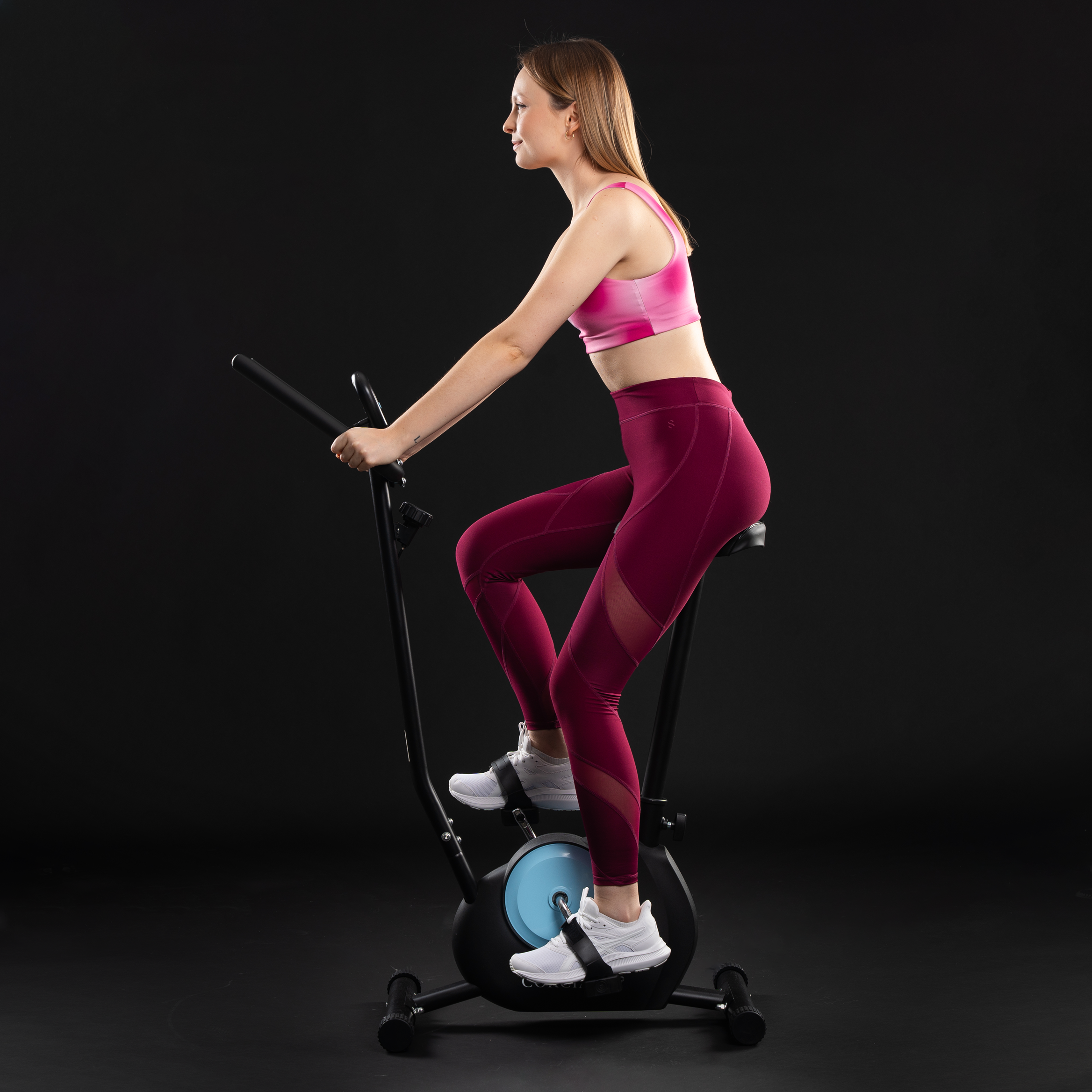 Kobieta w różowym topie sportowym i czerwonych legginsach jedzie na czarnym rowerku stacjonarnym na ciemnym tle.