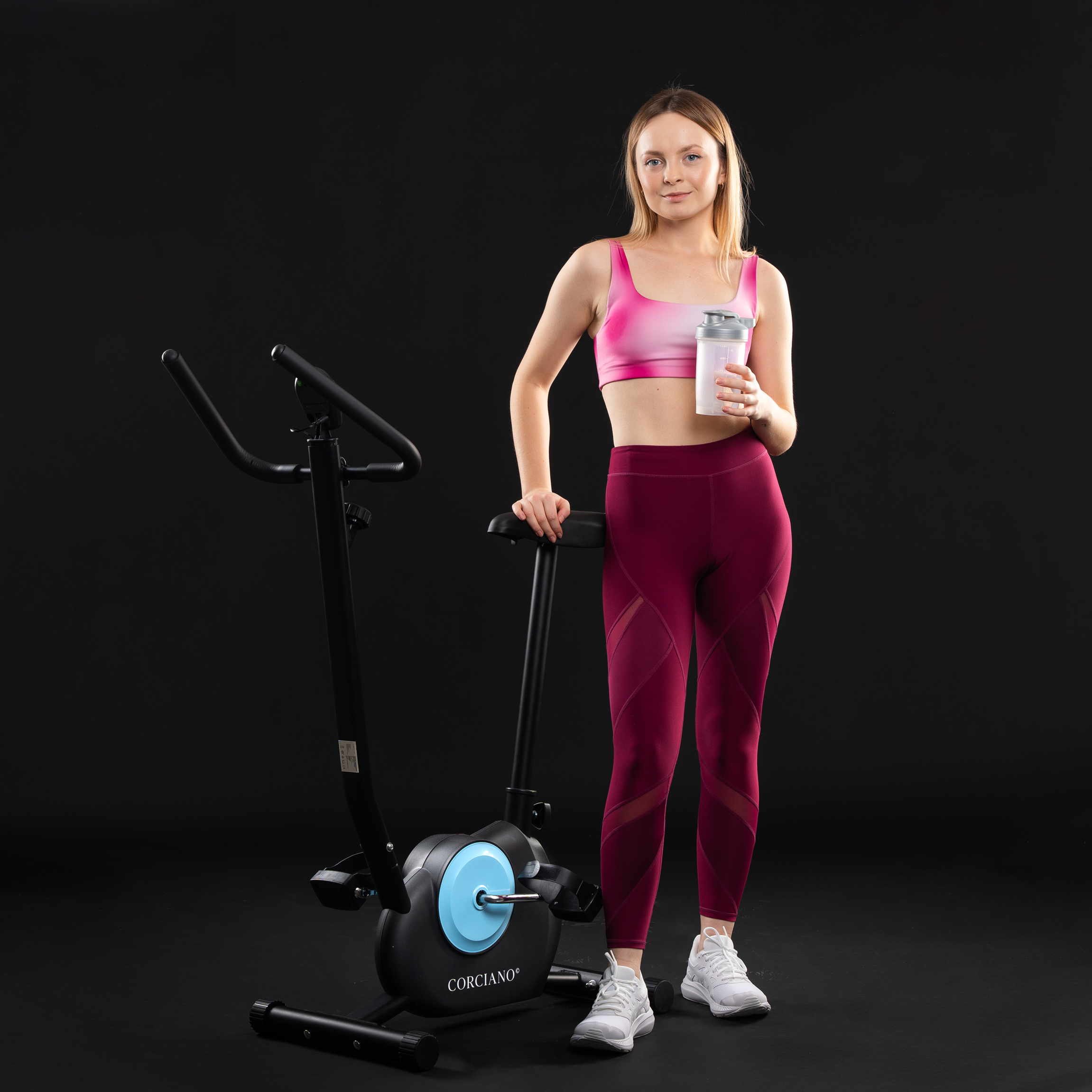 Blond kobieta w stroju sportowym stoi obok czarnego roweru stacjonarnego, trzymając szejker.