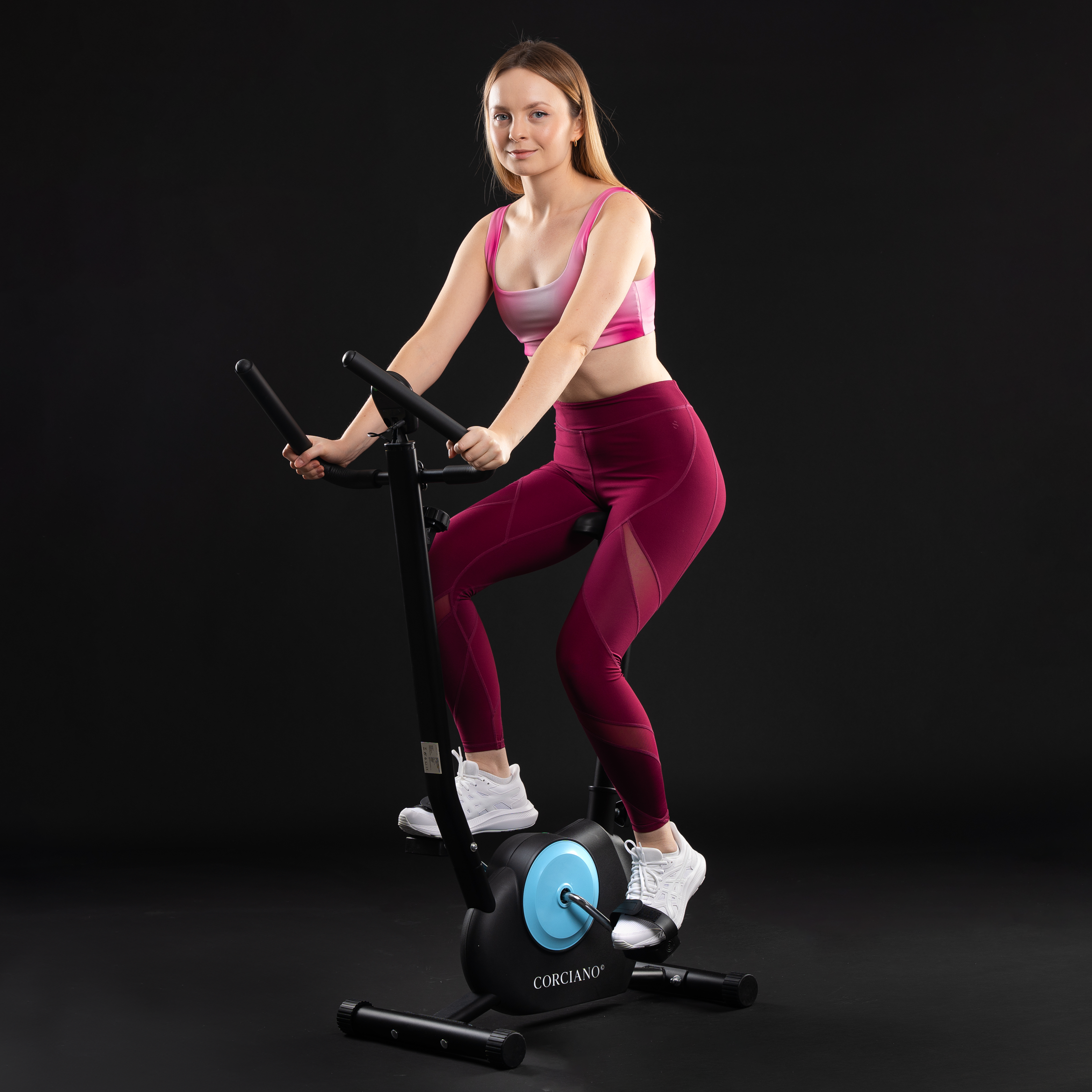 Blond kobieta w różowym stroju sportowym ćwiczy na rowerku stacjonarnym na czarnym tle.