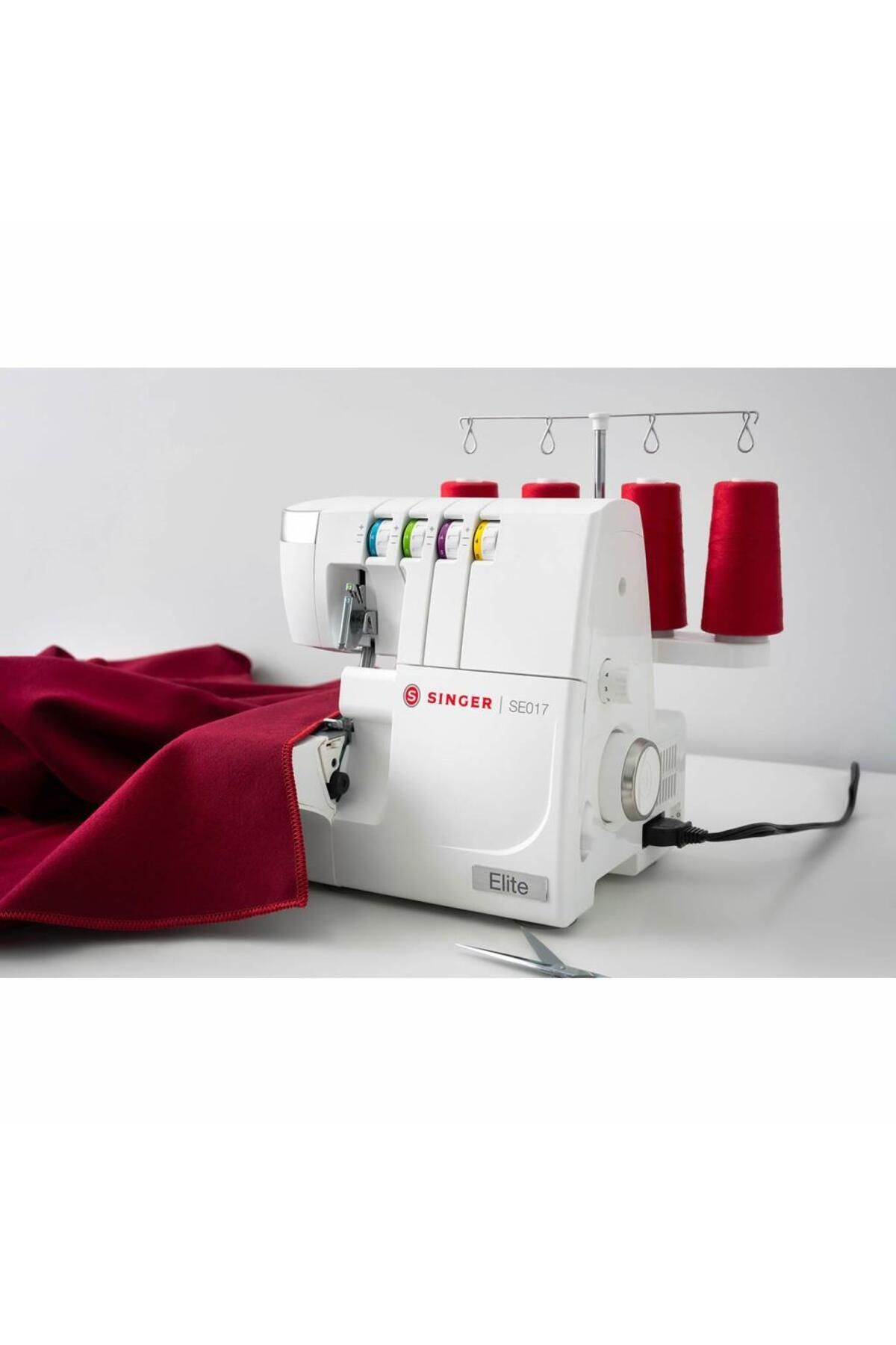 Kırmızı iplik makaraları ve kırmızı kumaş yanındaki beyaz Singer Elite SE017 overlok makinesi.