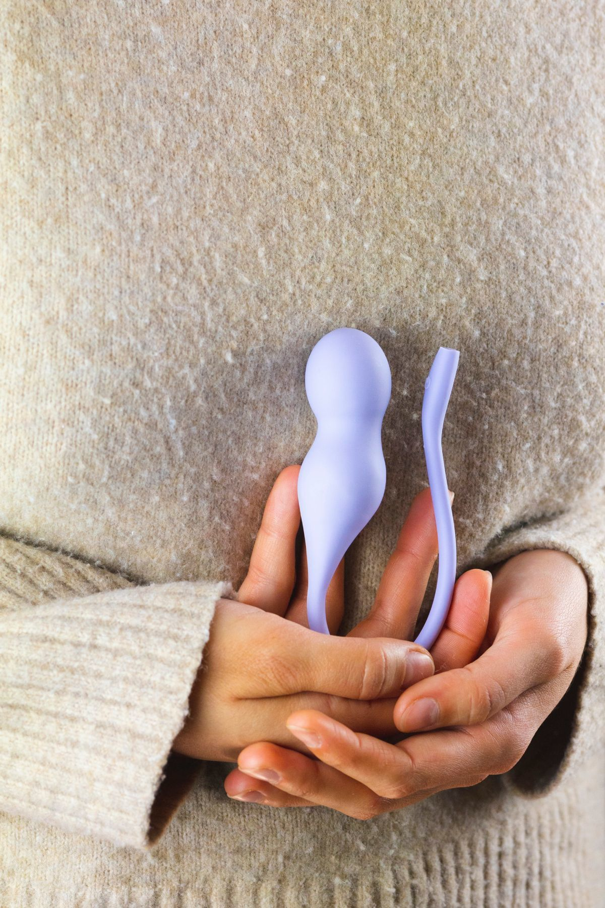Eine Person in einem beigen Pullover hält ein hellviolettes Wellnessprodukt aus Silikon an die Brust.