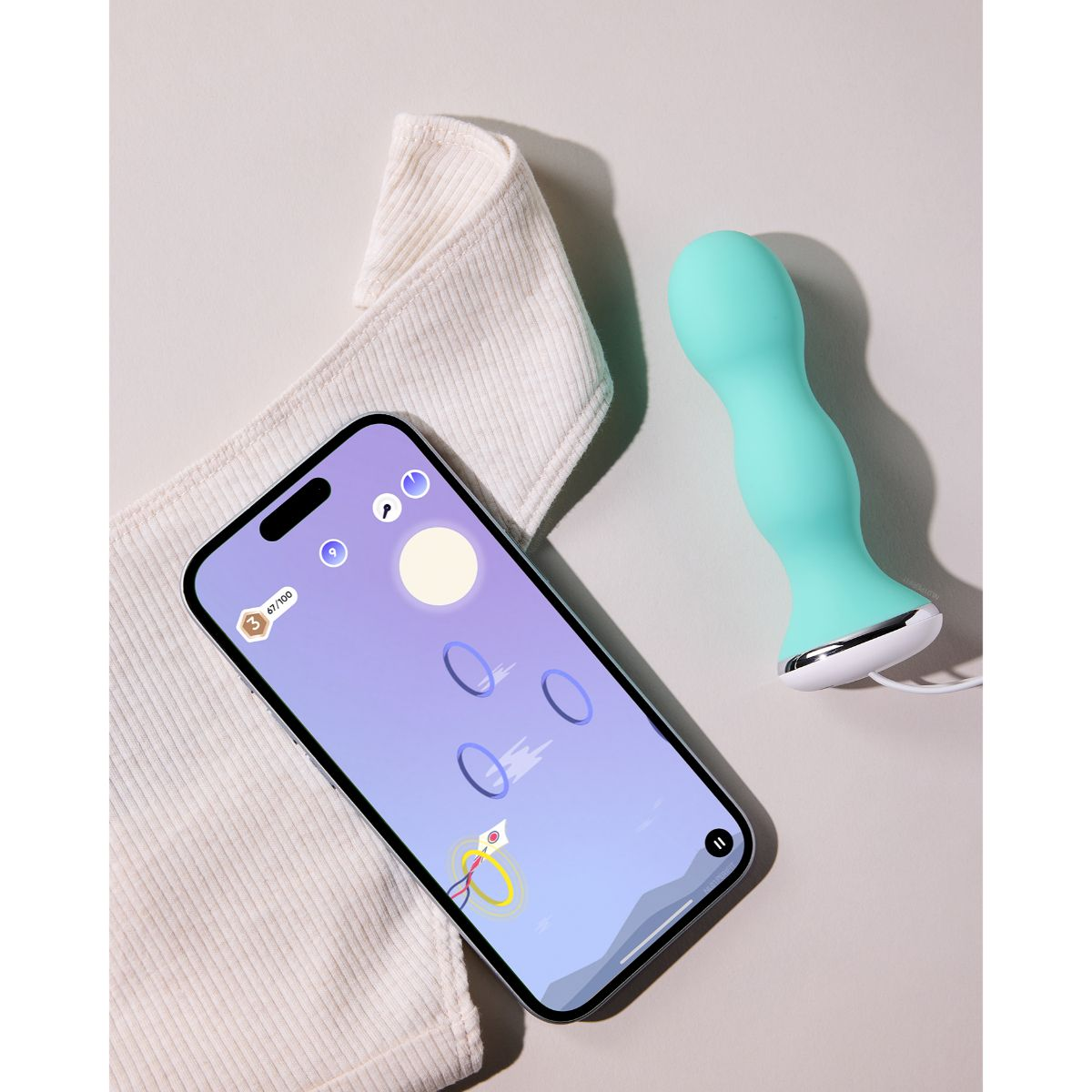 Ein Smartphone mit App-Spiel, ein beiges Textil und ein hellblaues, kurviges Gerät auf beigem Untergrund.