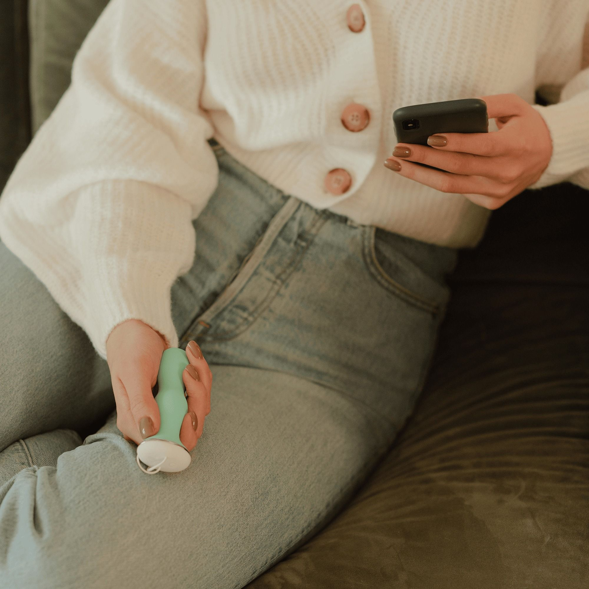 Eine Frau in weißem Cardigan und Jeans hält ein Smartphone und ein türkises Gadget auf einem Sofa.