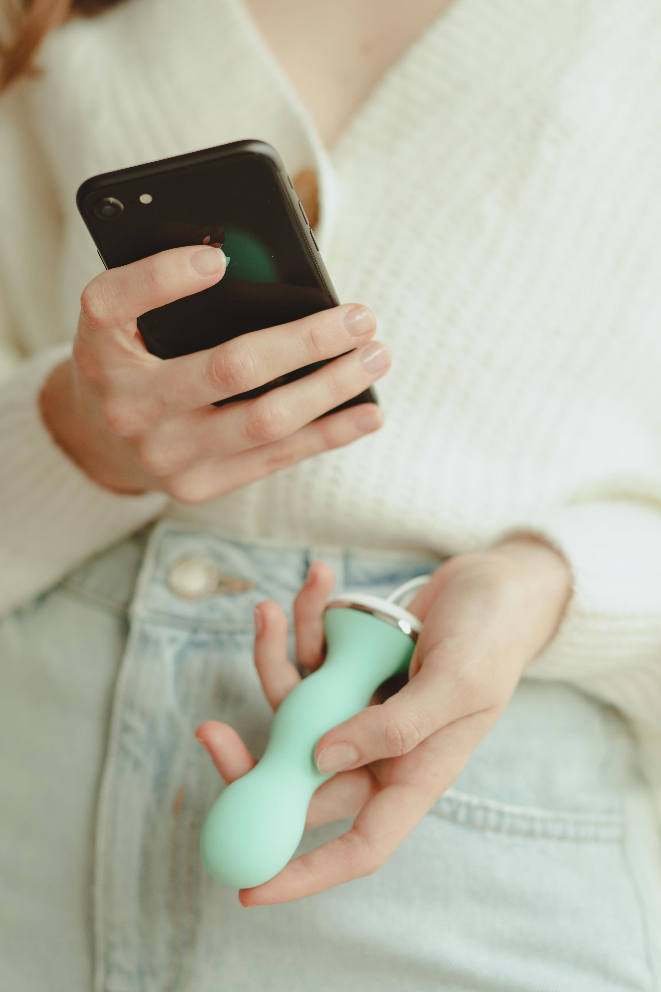 Eine Frau im weißen Pullover hält ein schwarzes Smartphone in der einen und ein mintgrünes Sexspielzeug in der anderen Hand.