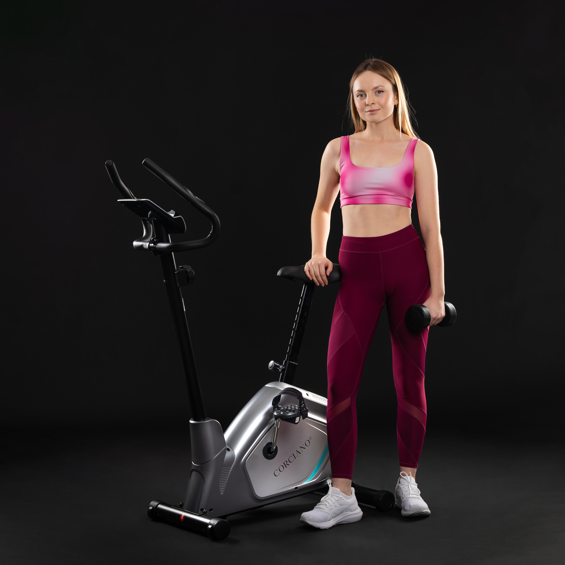 Kobieta w stroju sportowym stoi obok roweru stacjonarnego, trzymając hantel.