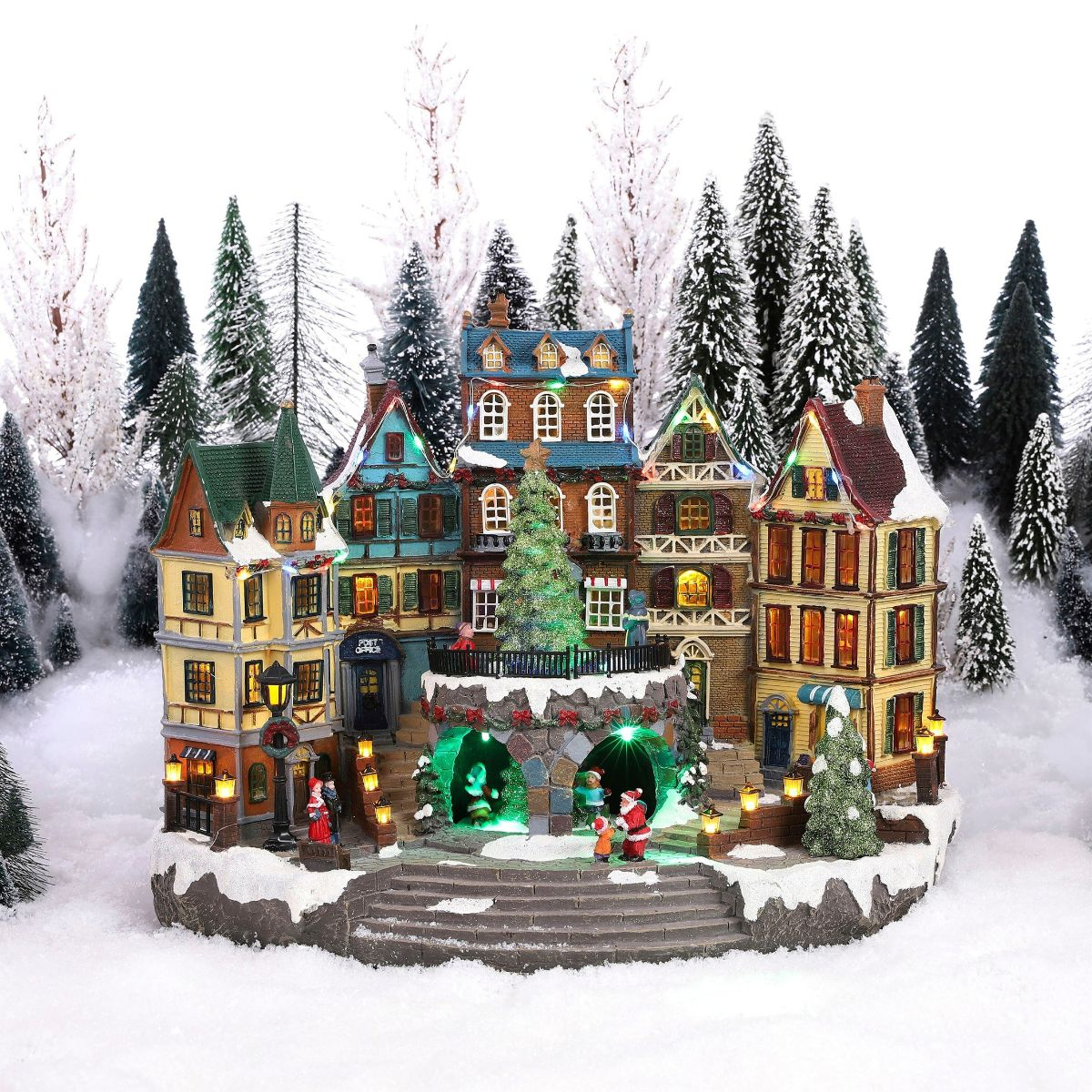 Village de Noël illuminé dans la neige avec maisons colorées, sapin de Noël et figurines devant des sapins enneigés.