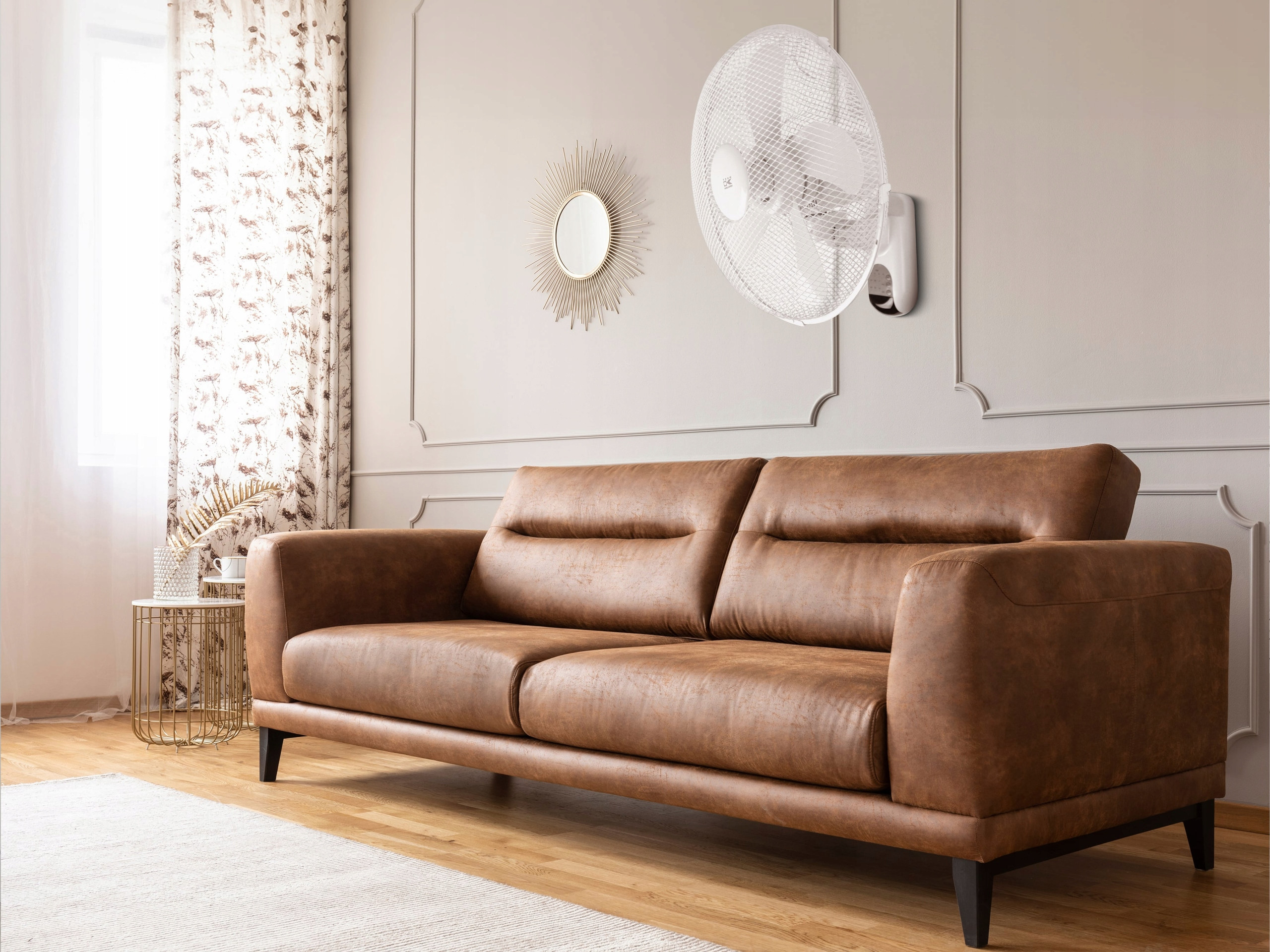 Brunatna skórzana sofa stoi w salonie z szarą ścianą, ozdobnym lustrem i białym wentylatorem ściennym.