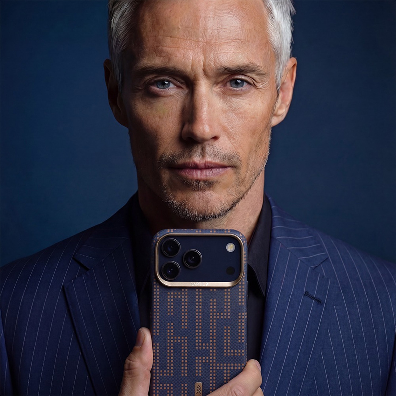 Un hombre con canas y traje azul sostiene un smartphone con funda estampada y varias cámaras frente a él.