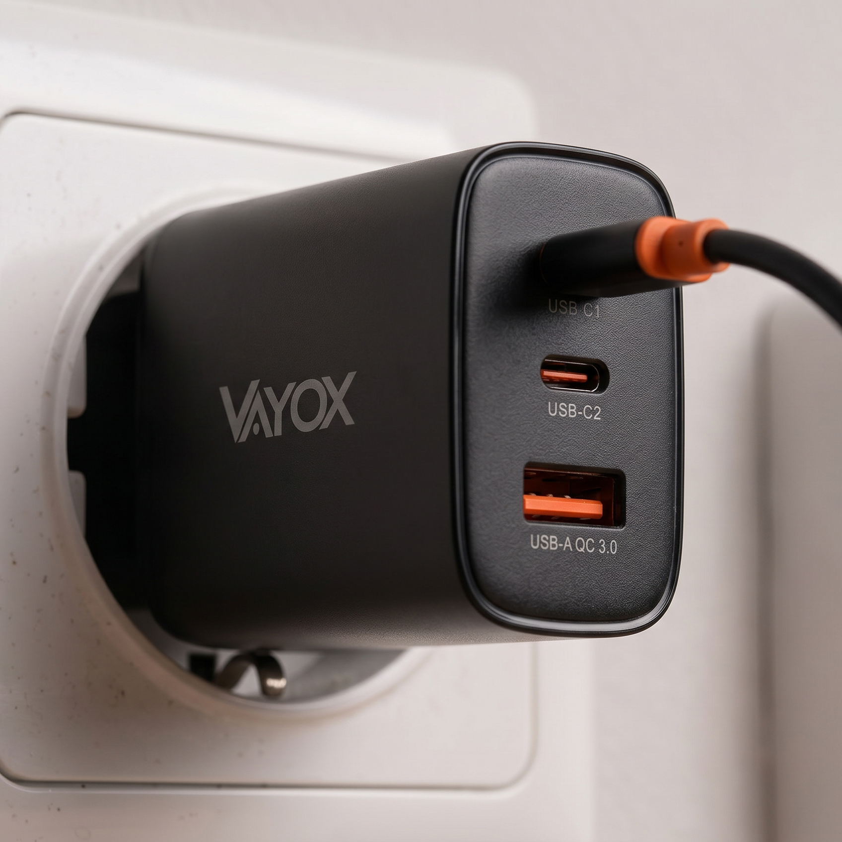 Egy fekete Vayox fali töltő fehér aljzatba dugva, csatlakoztatott USB-kábellel.