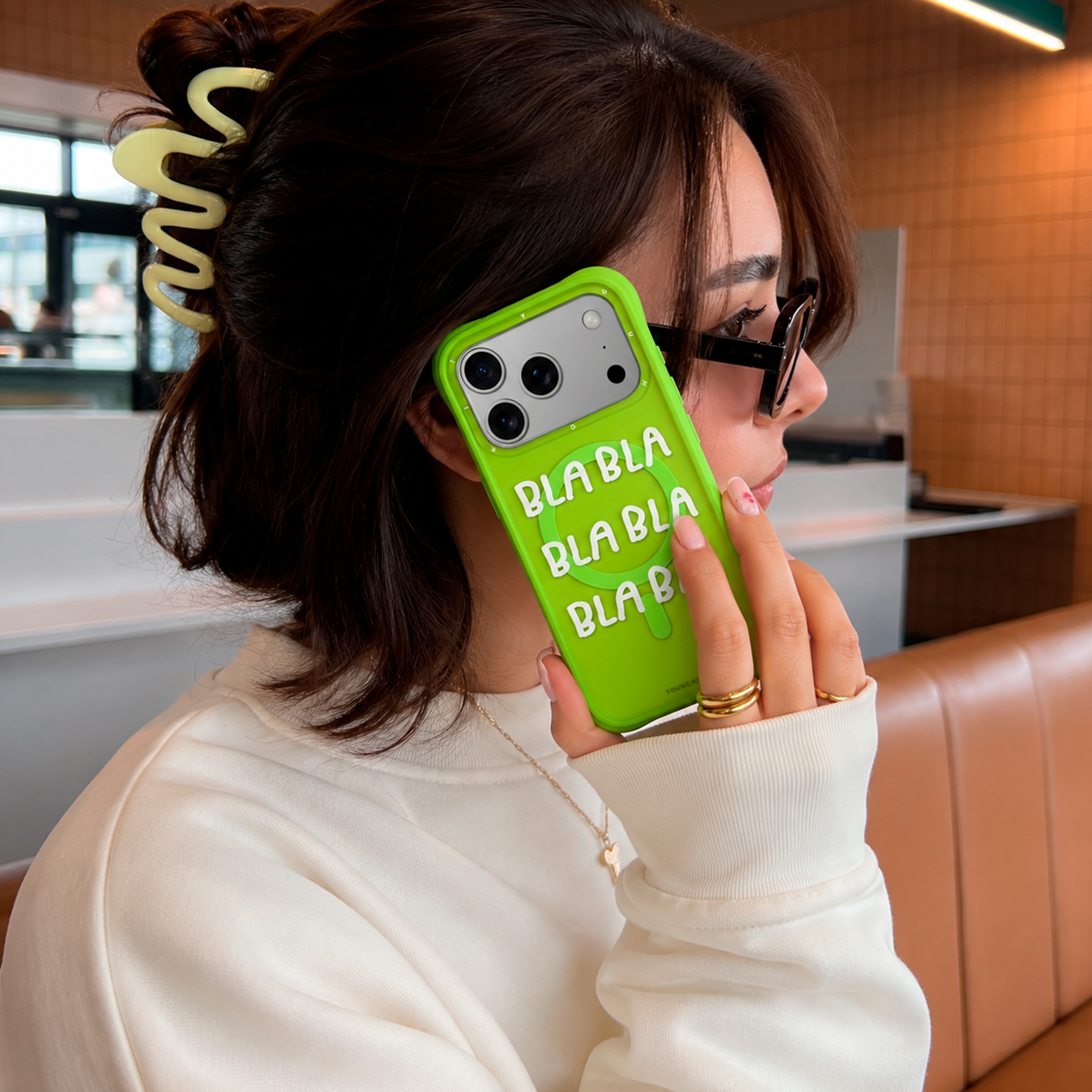 Una mujer sostiene una funda de teléfono verde brillante con texto blanco "BLA BLA", vistiendo un suéter crema y una pinza.