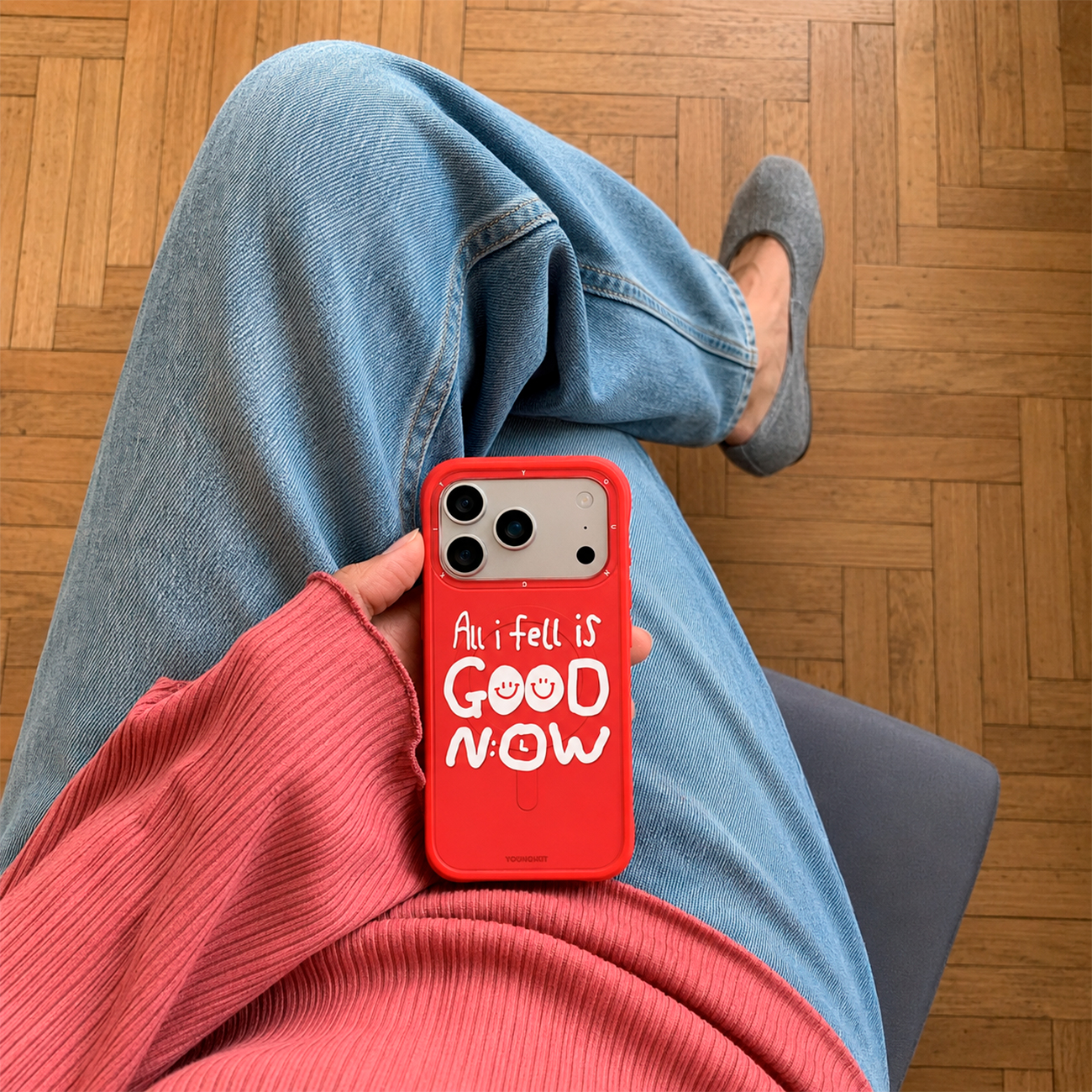 Persona en jeans y top rosa sostiene un teléfono rojo con texto blanco sobre suelo de madera.