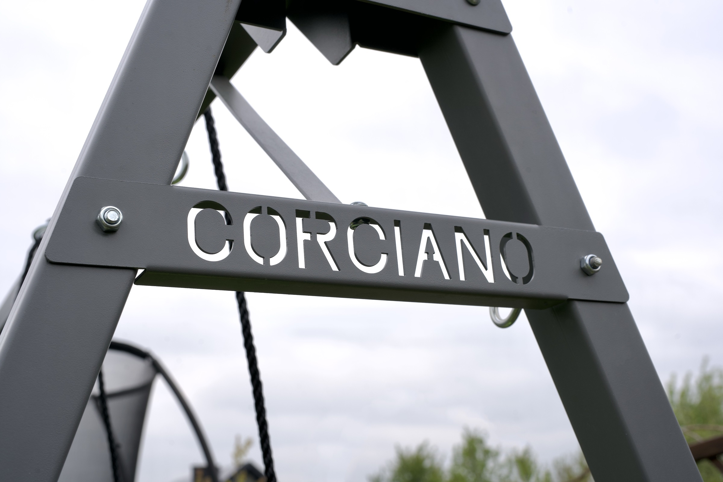 Szara metalowa konstrukcja huśtawki z wyciętym napisem CORCIANO na tle pochmurnego nieba.