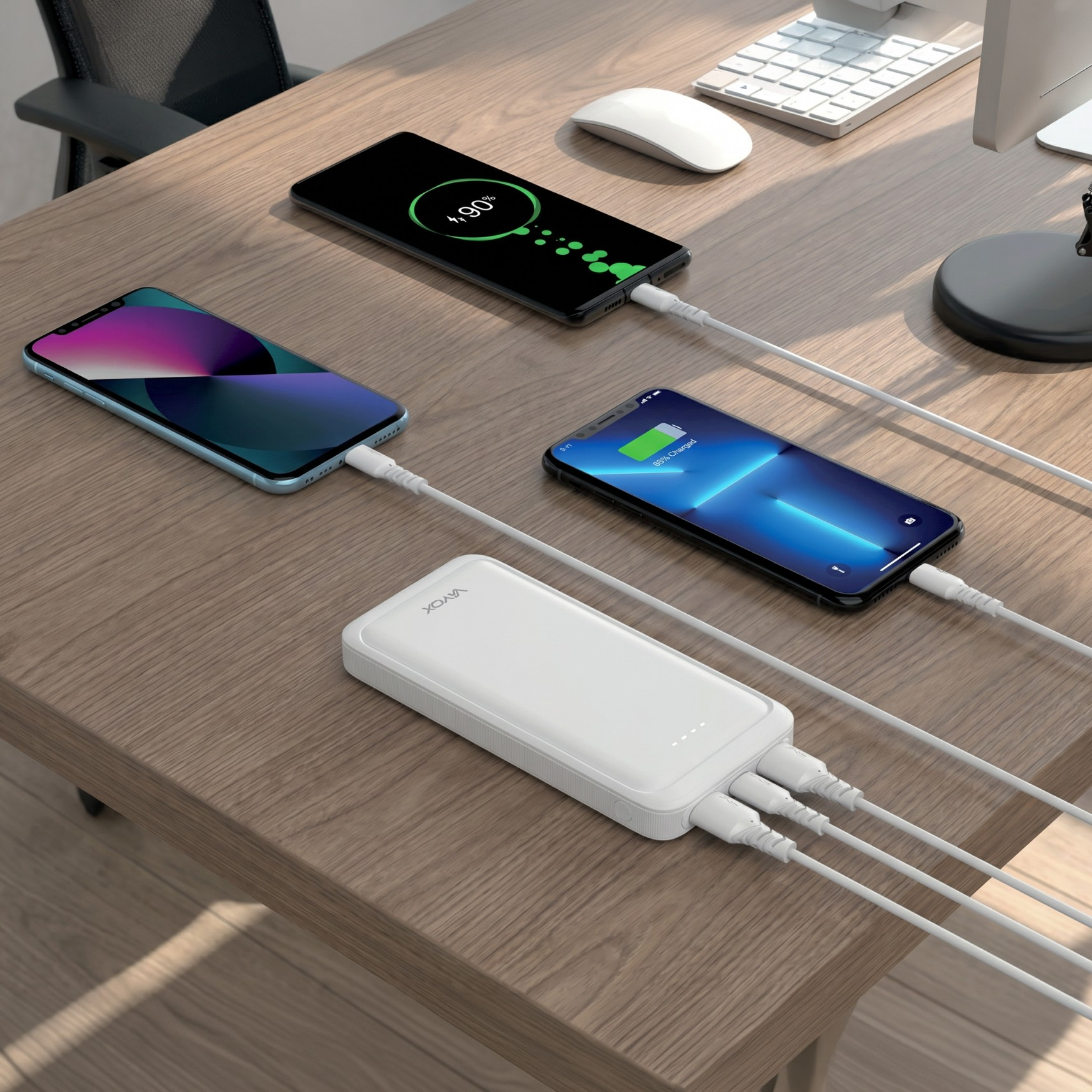 Biały powerbank ładuje trzy smartfony na drewnianym biurku obok klawiatury.