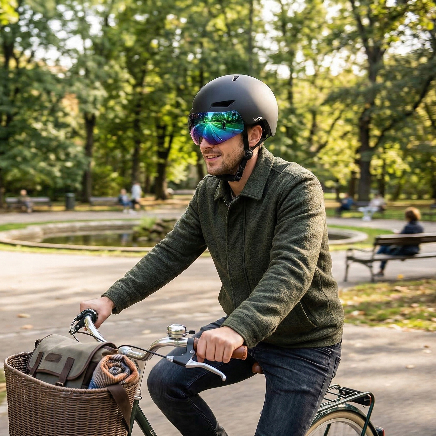 Mężczyzna w zielonej kurtce i czarnym kasku z wizjerem jedzie na rowerze przez park z zielonymi drzewami.