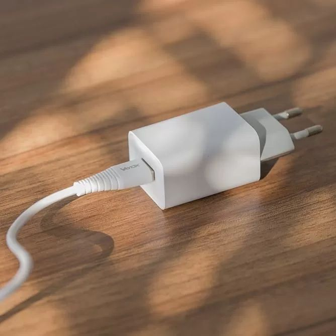 Egy fehér USB fali töltő és kábel fekszik egy fa felületen a napfényben.