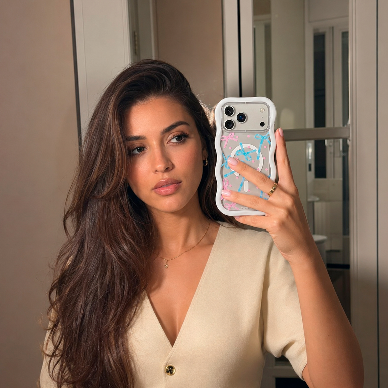 Una mujer con cabello castaño largo y un top crema se toma una selfi frente al espejo.