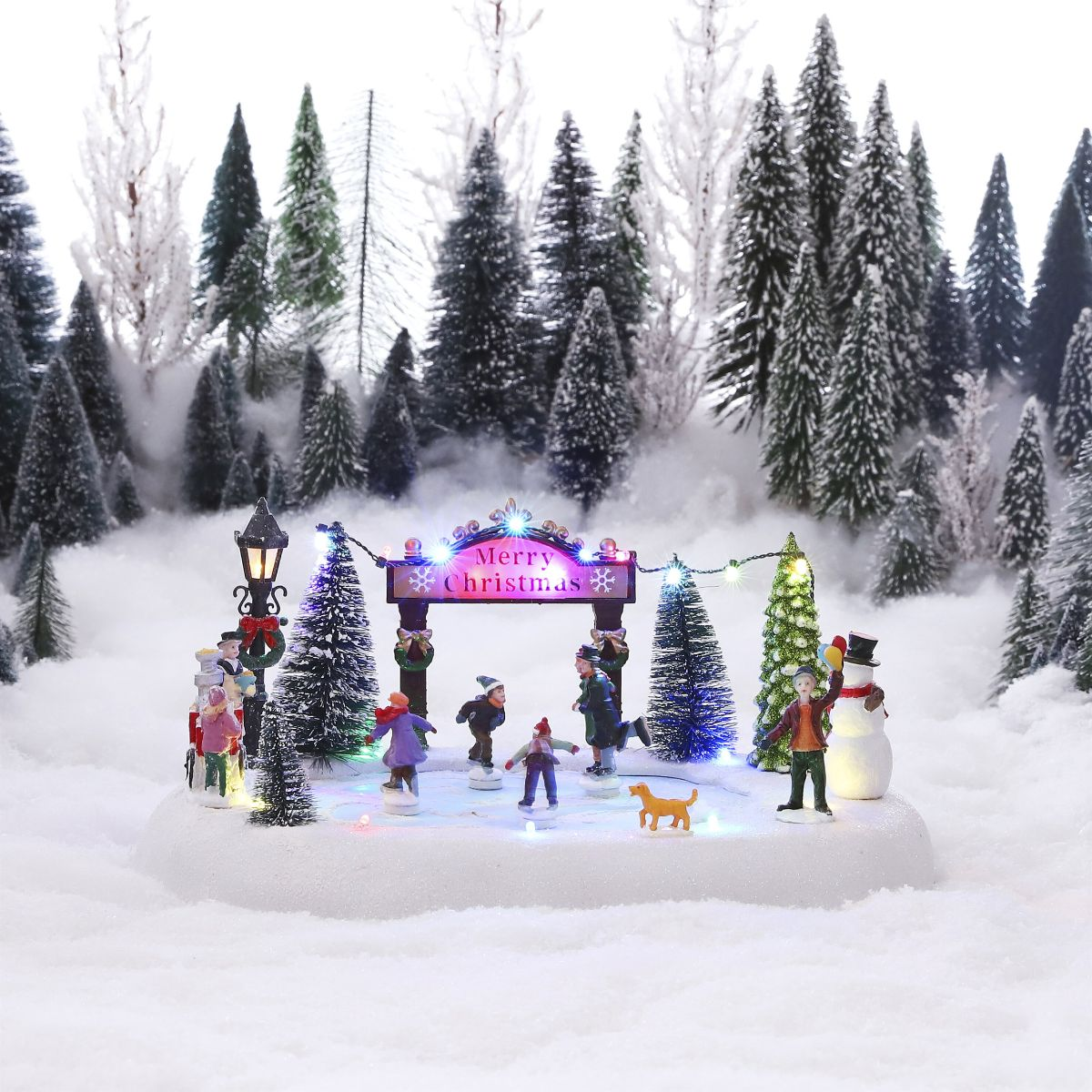 Scène de Noël miniature avec patineurs, sapins, panneau Joyeux Noël et paysage enneigé en arrière-plan.