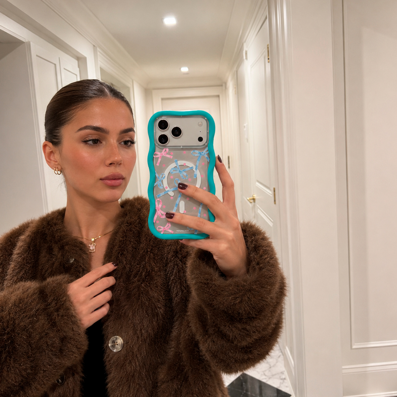 Una mujer con abrigo de piel marrón se hace un selfie frente al espejo con un móvil con funda turquesa.