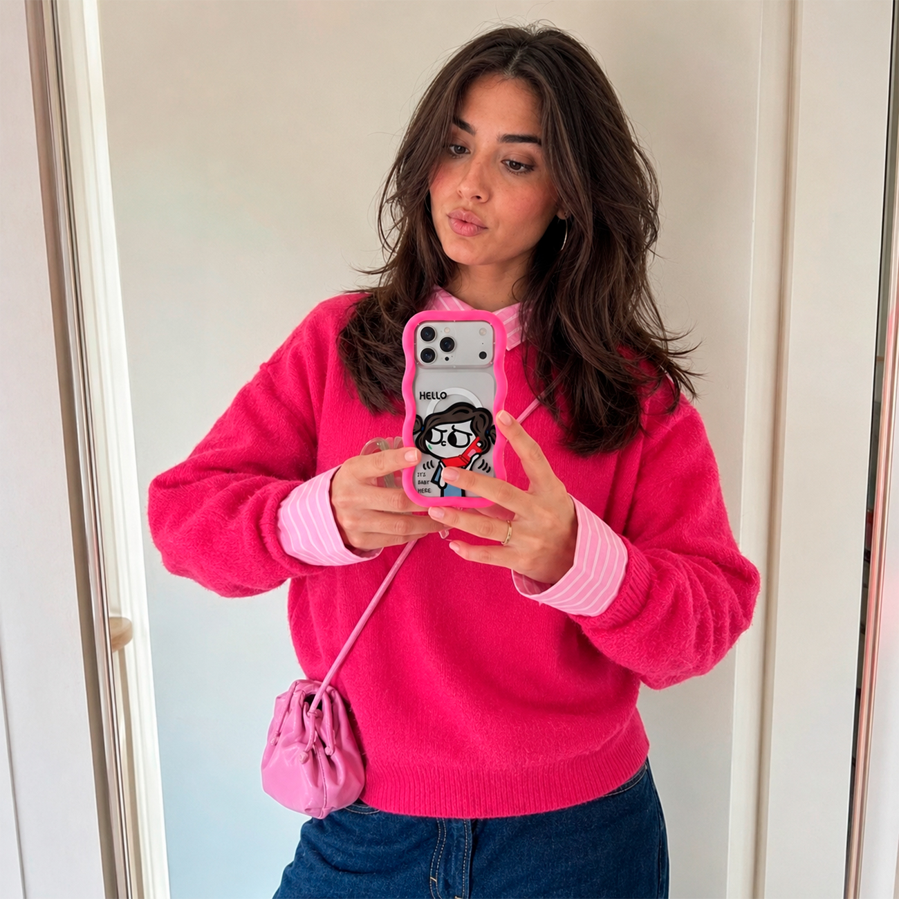 Una mujer con cabello oscuro, jersey rosa y jeans azules sostiene un teléfono con funda rosa para un selfi.