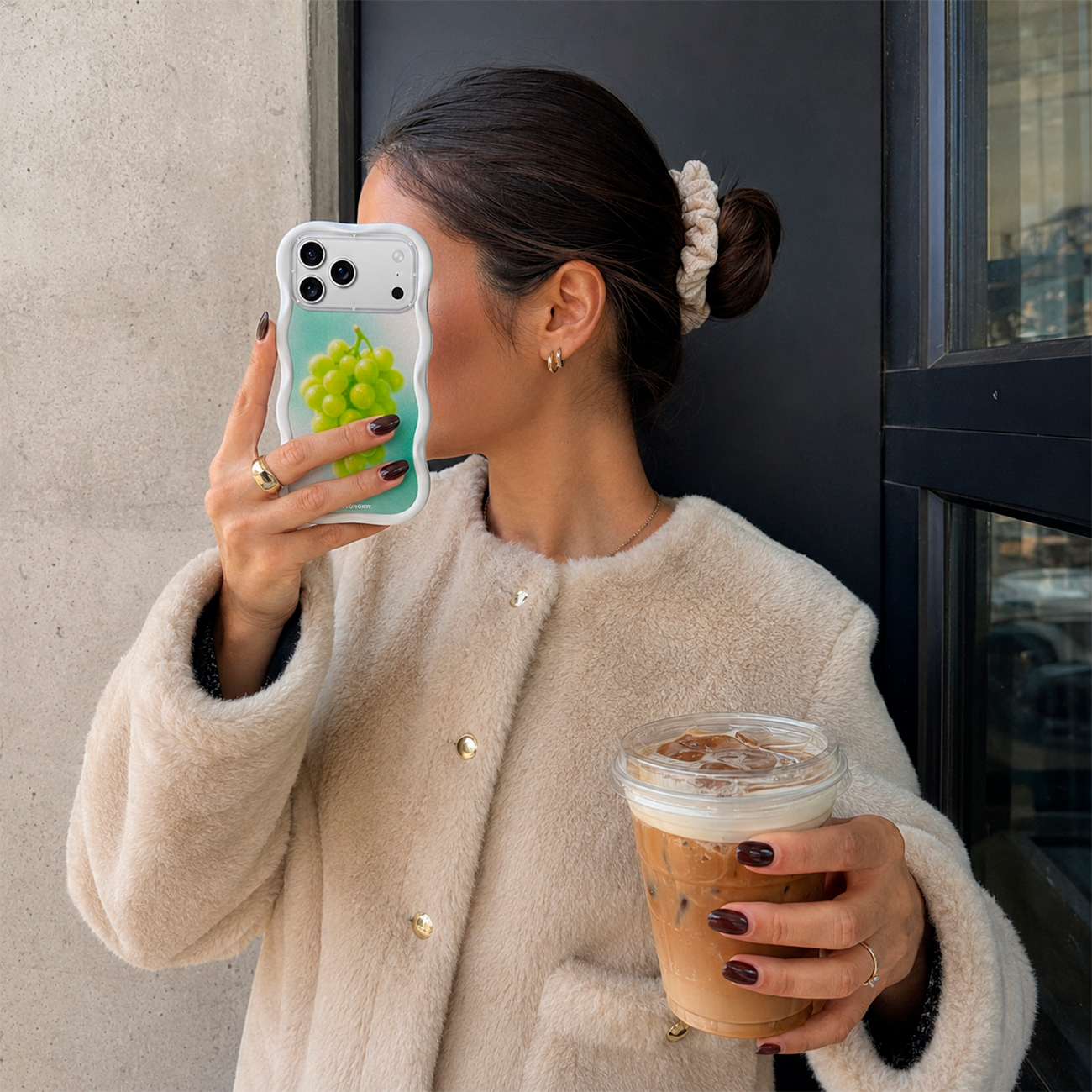 Una mujer con abrigo polar beis sostiene un café con hielo y toma una foto con una funda de móvil de uvas.