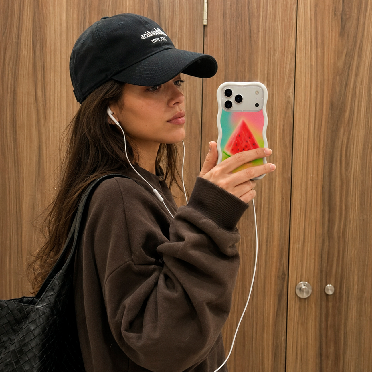 Una mujer con gorra negra y suéter marrón se toma un selfi en el espejo con un teléfono con patrón de sandía.