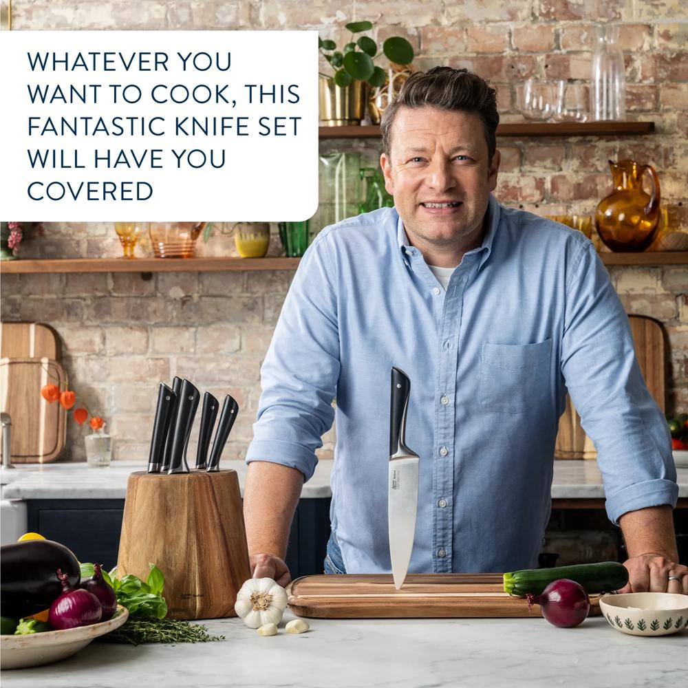 Jamie Oliver lächelt in einer Küche neben einem Holzblock mit Messern und Gemüse auf der Arbeitsplatte.