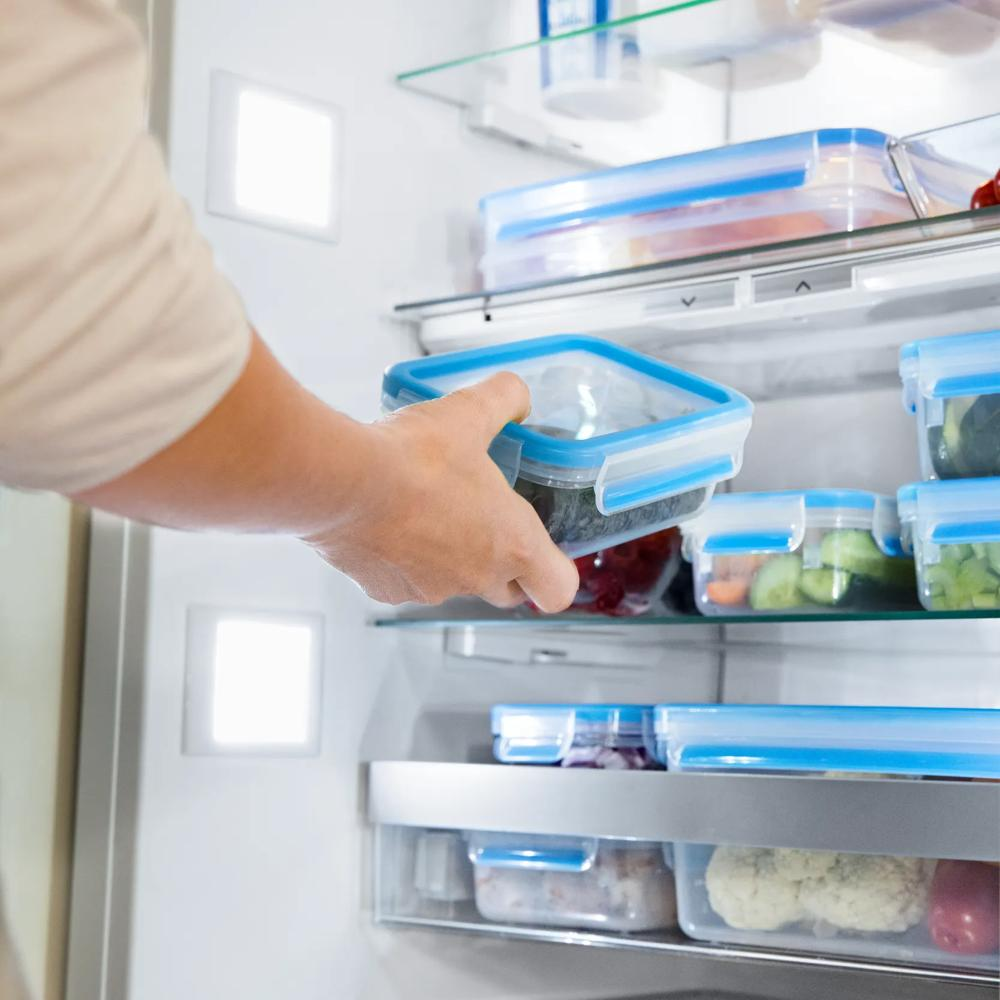 Person nimmt eine quadratische Plastikdose mit blauem Deckel aus einem gut gefüllten, beleuchteten Kühlschrank.