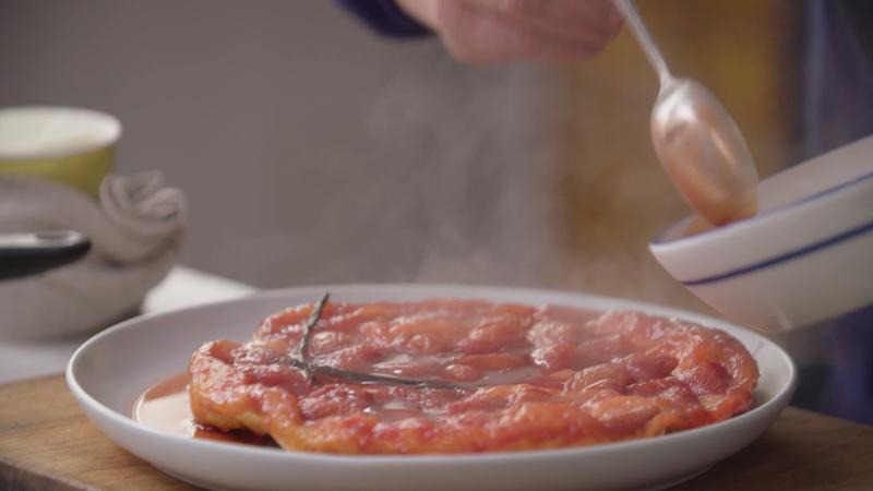 Person gießt Sauce mit Löffel auf eine warme Obsttorte mit Vanilleschote auf weißem Teller.