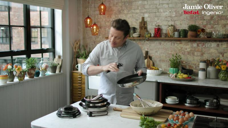 Koch Jamie Oliver hält eine Bratpfanne in einer hellen Küche mit Ziegelwänden, Kochgeschirr und Fenster.