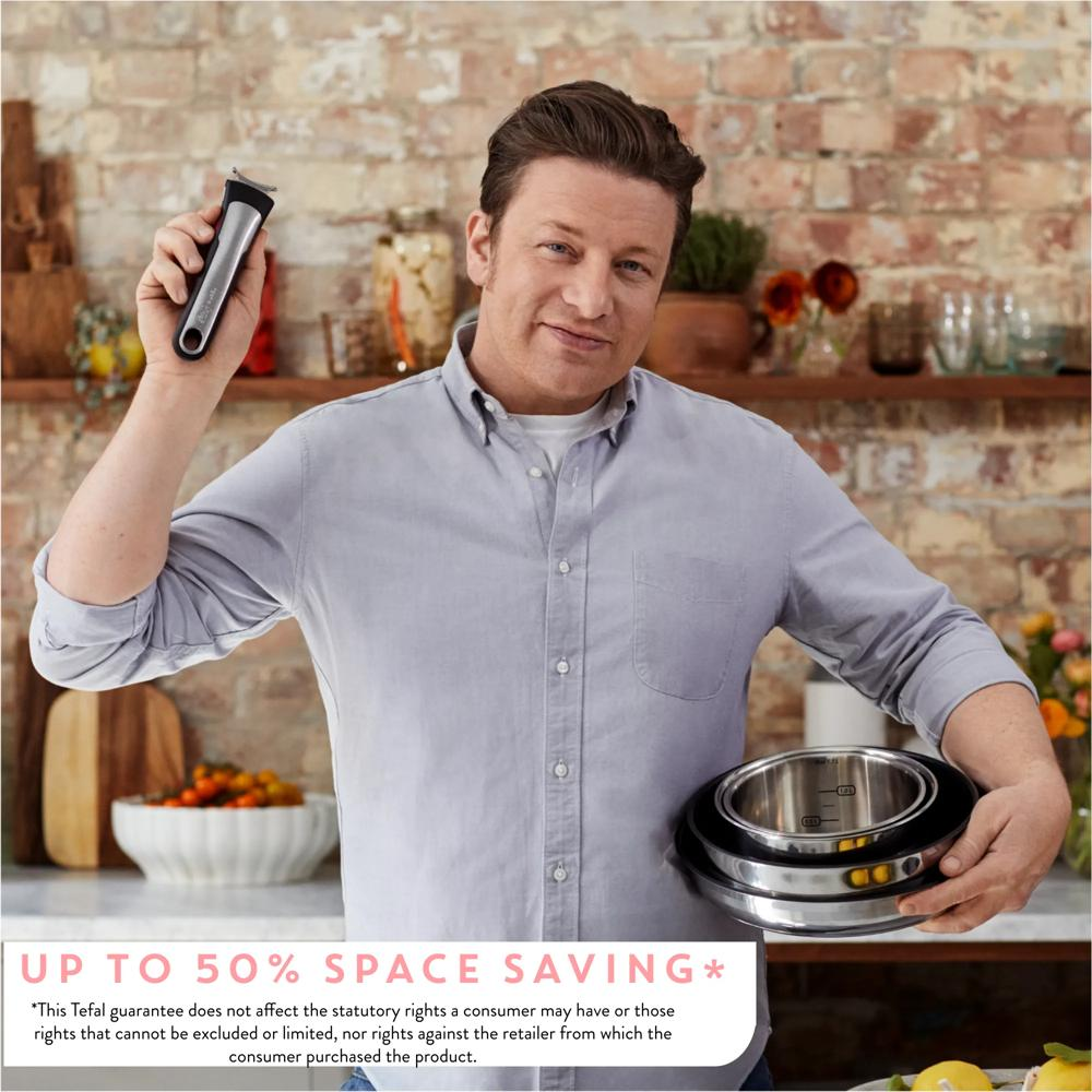 Jamie Oliver hält in einer Küche einen abnehmbaren Griff und einen Stapel Töpfe vor einer Ziegelwand.
