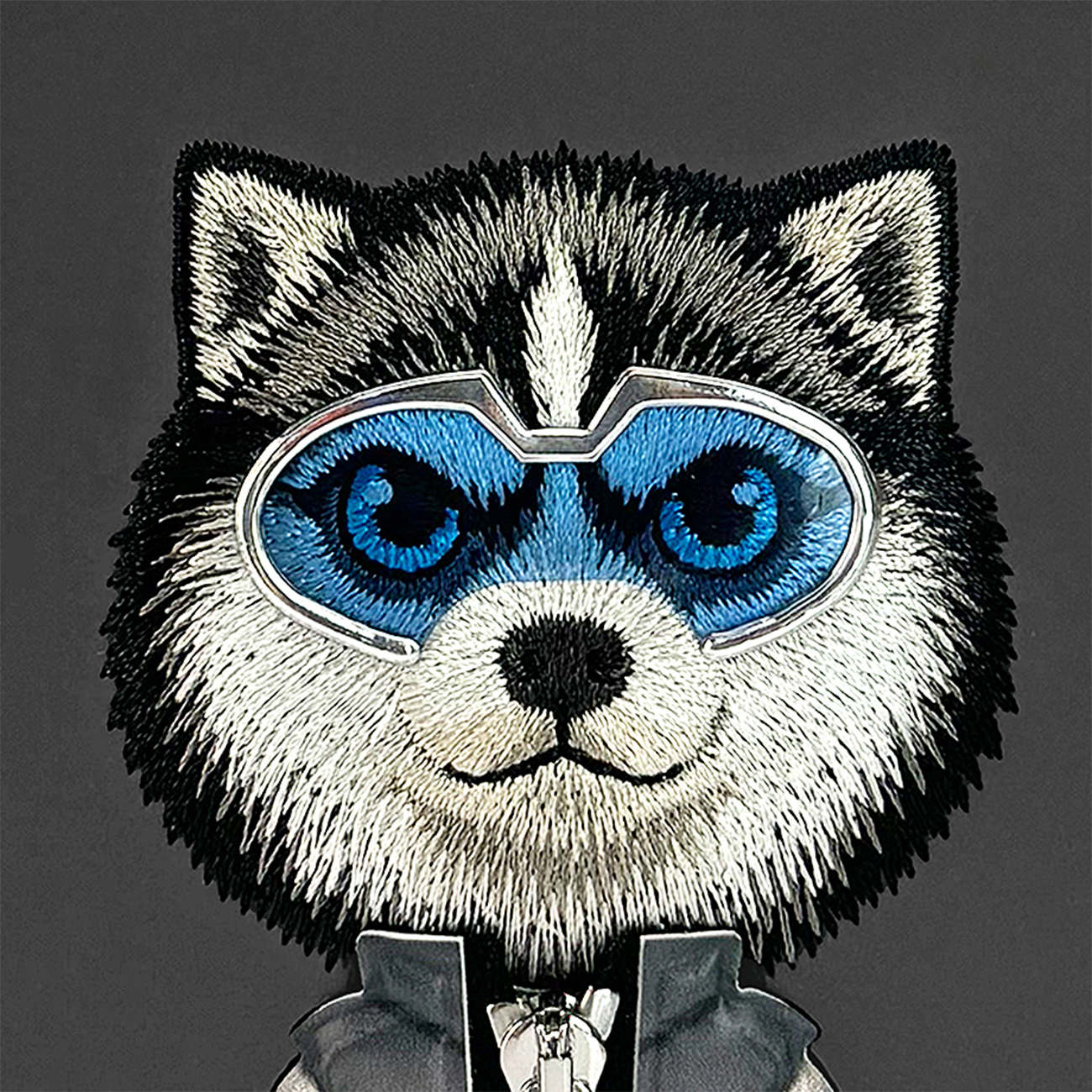 Imagen bordada de una cabeza de lobo con ojos azules, máscara azul y una cremallera sobre fondo gris.