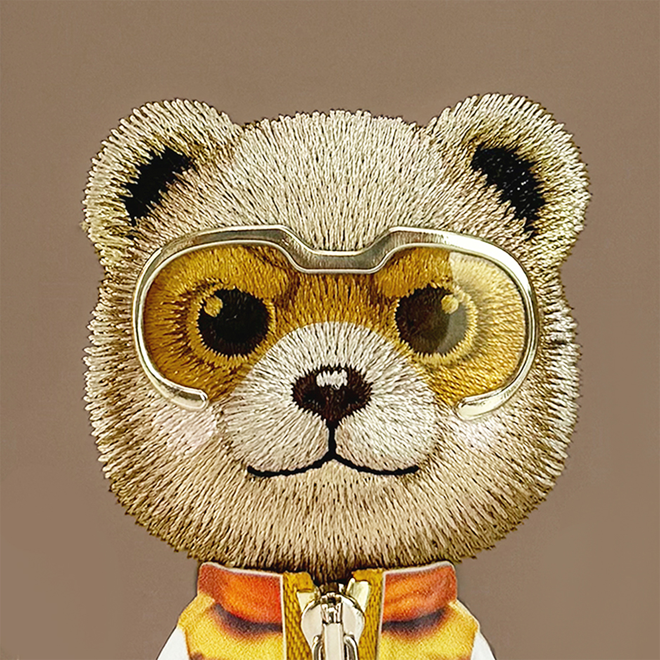 Cara de oso de peluche bordada con gafas de esquí y chaqueta de cuello naranja sobre fondo sólido.
