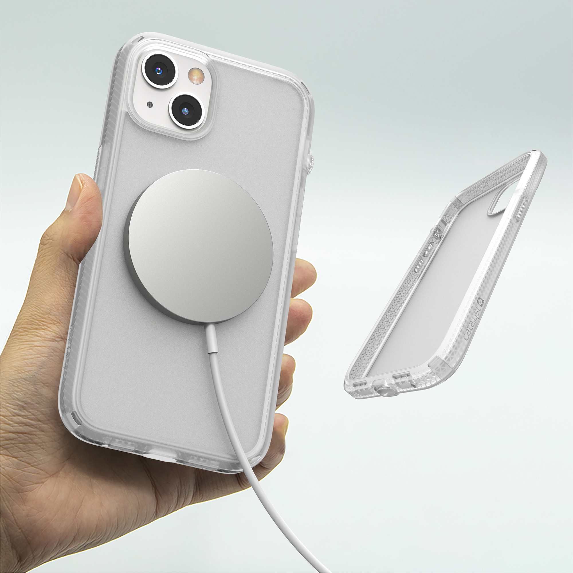 Hand hält weißes Smartphone mit MagSafe-Ladegerät, daneben eine transparente Schutzhülle auf hellem Hintergrund.