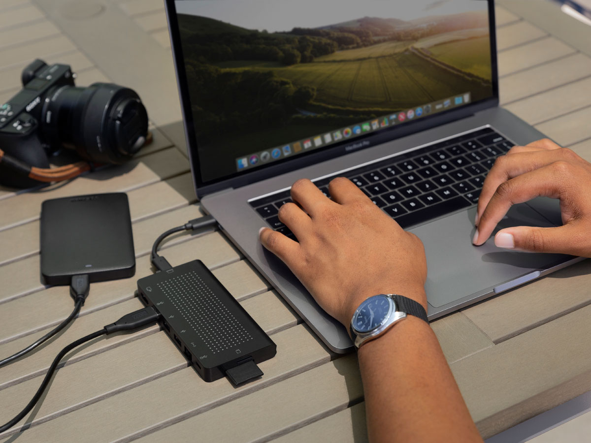 Person tippt auf einem Laptop, verbunden mit einem USB-Hub. Eine Kamera und Festplatte liegen auf dem Holztisch.