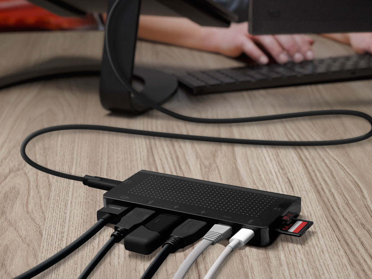 Ein schwarzer USB-Hub mit angeschlossenen Kabeln und SD-Karten auf einem Holzschreibtisch, im Hintergrund tippt jemand.