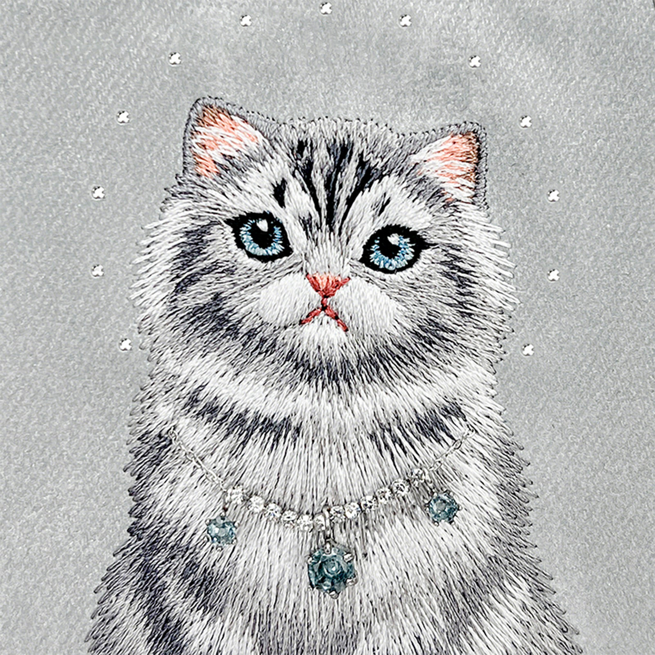 Gato gris bordado con ojos azules y un collar brillante sobre fondo gris.
