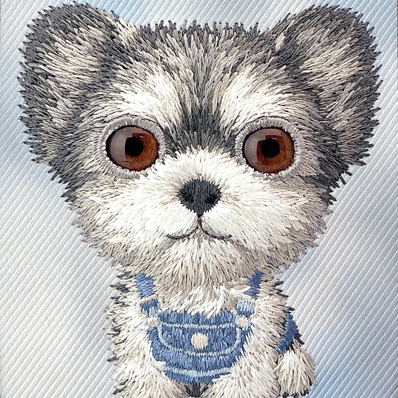 Imagen bordada de un animal esponjoso gris y blanco con grandes ojos marrones y peto azul.