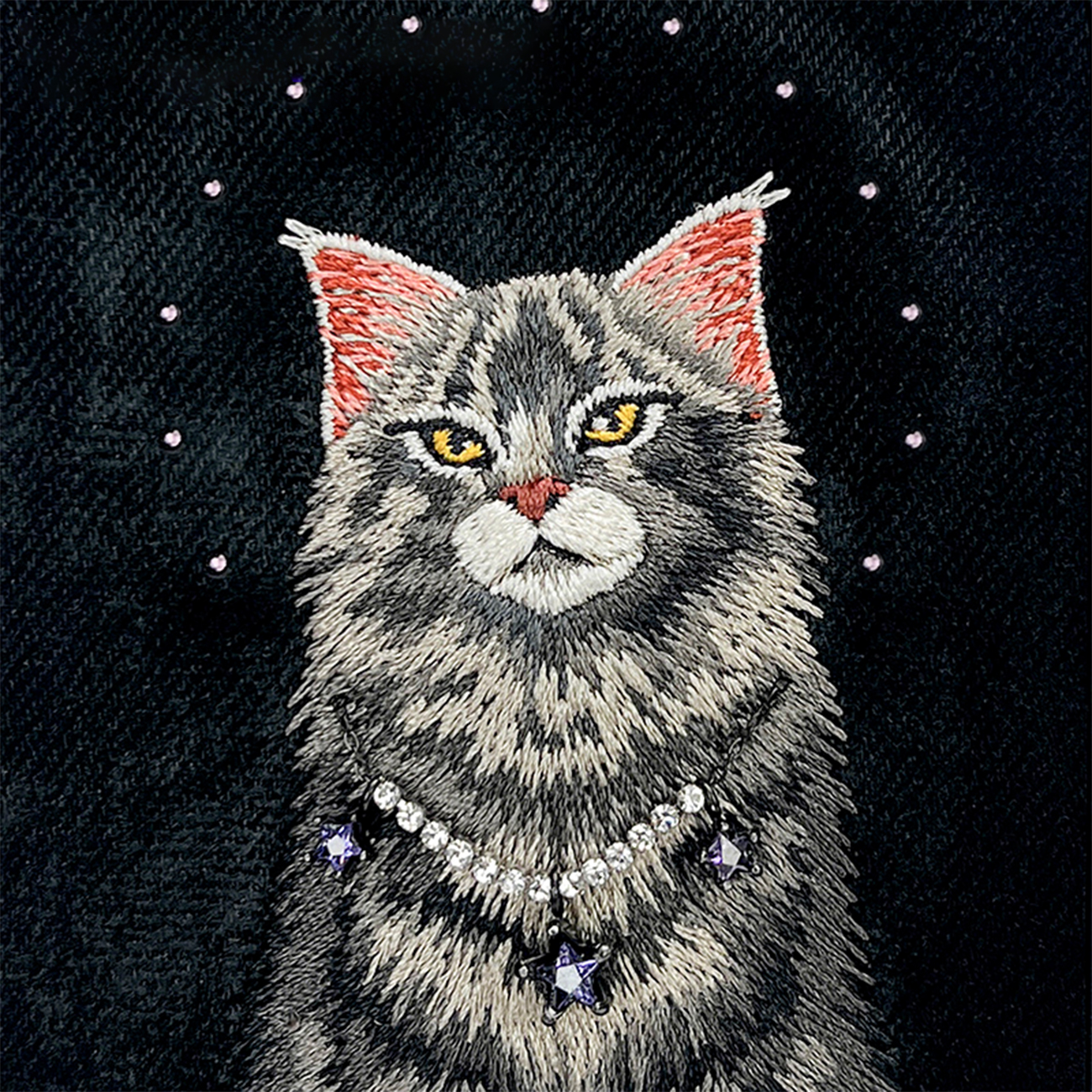 Retrato bordado de un gato gris con ojos amarillos y un collar de estrellas brillante sobre tela negra.