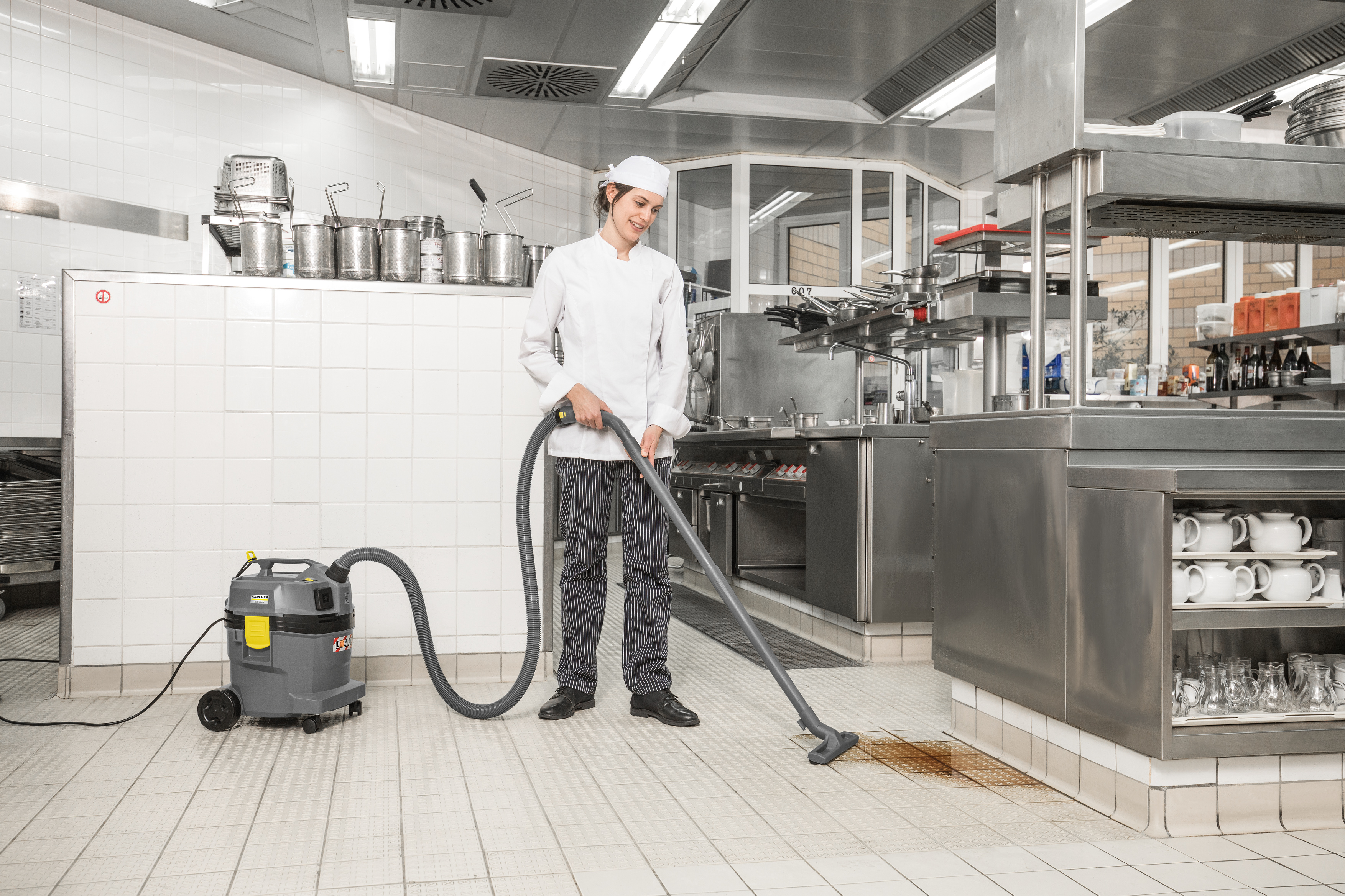 Uno chef in uniforme bianca pulisce il pavimento di una cucina commerciale con un aspirapolvere grigio.
