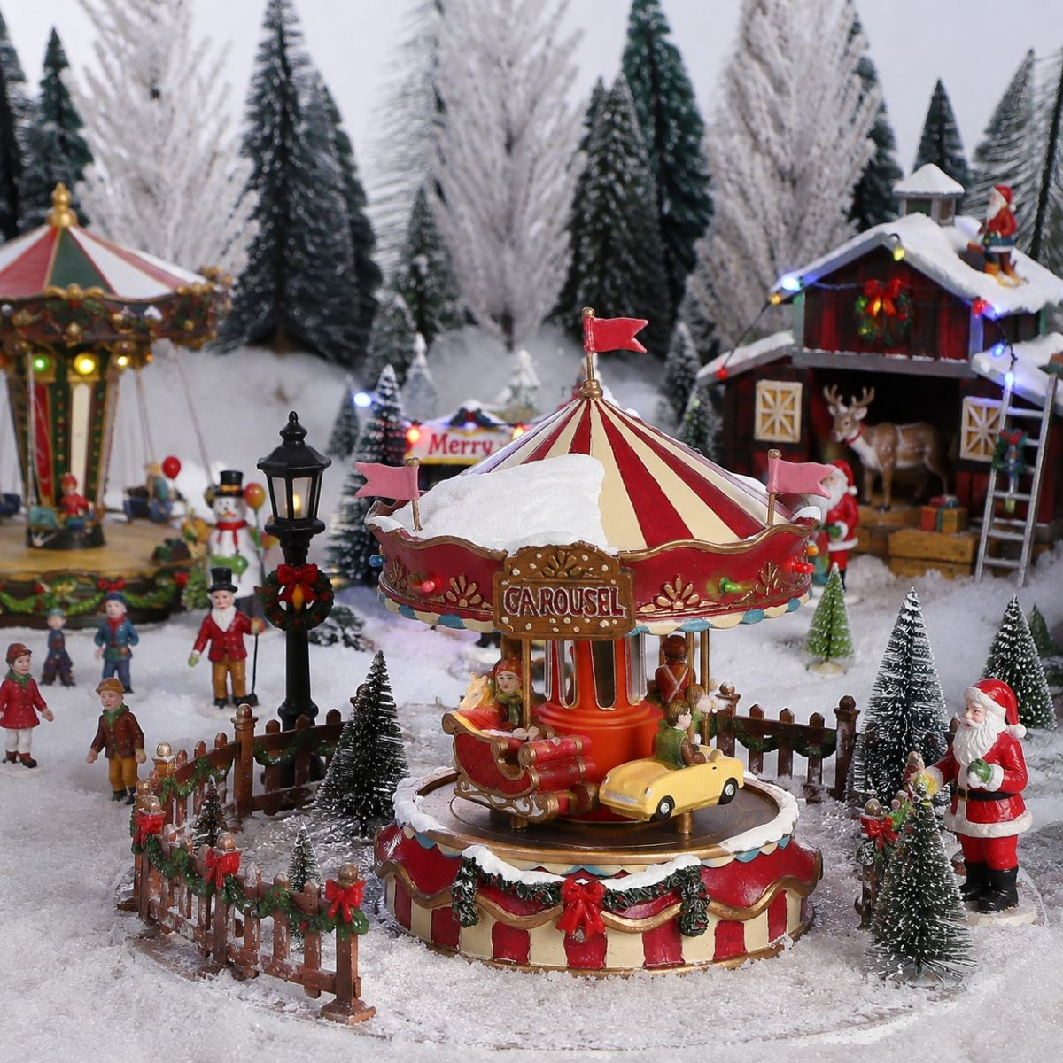 Een feestelijke kerstcarrousel-miniatuur in de sneeuw, omringd door bomen, figuren en een verlichte hut.