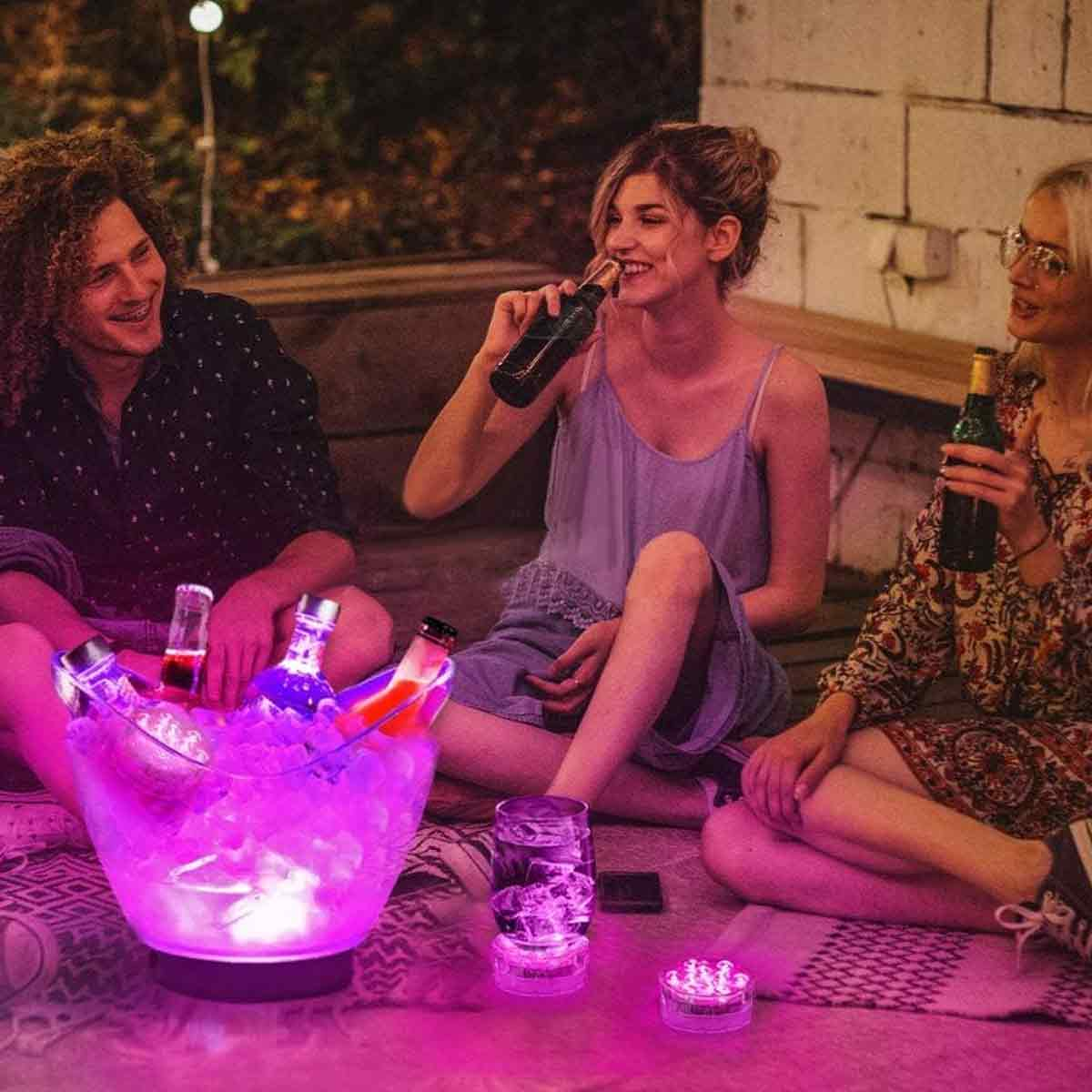 Freunde sitzen nachts draußen, trinken und lachen neben einem leuchtenden Eiswürfelbehälter.