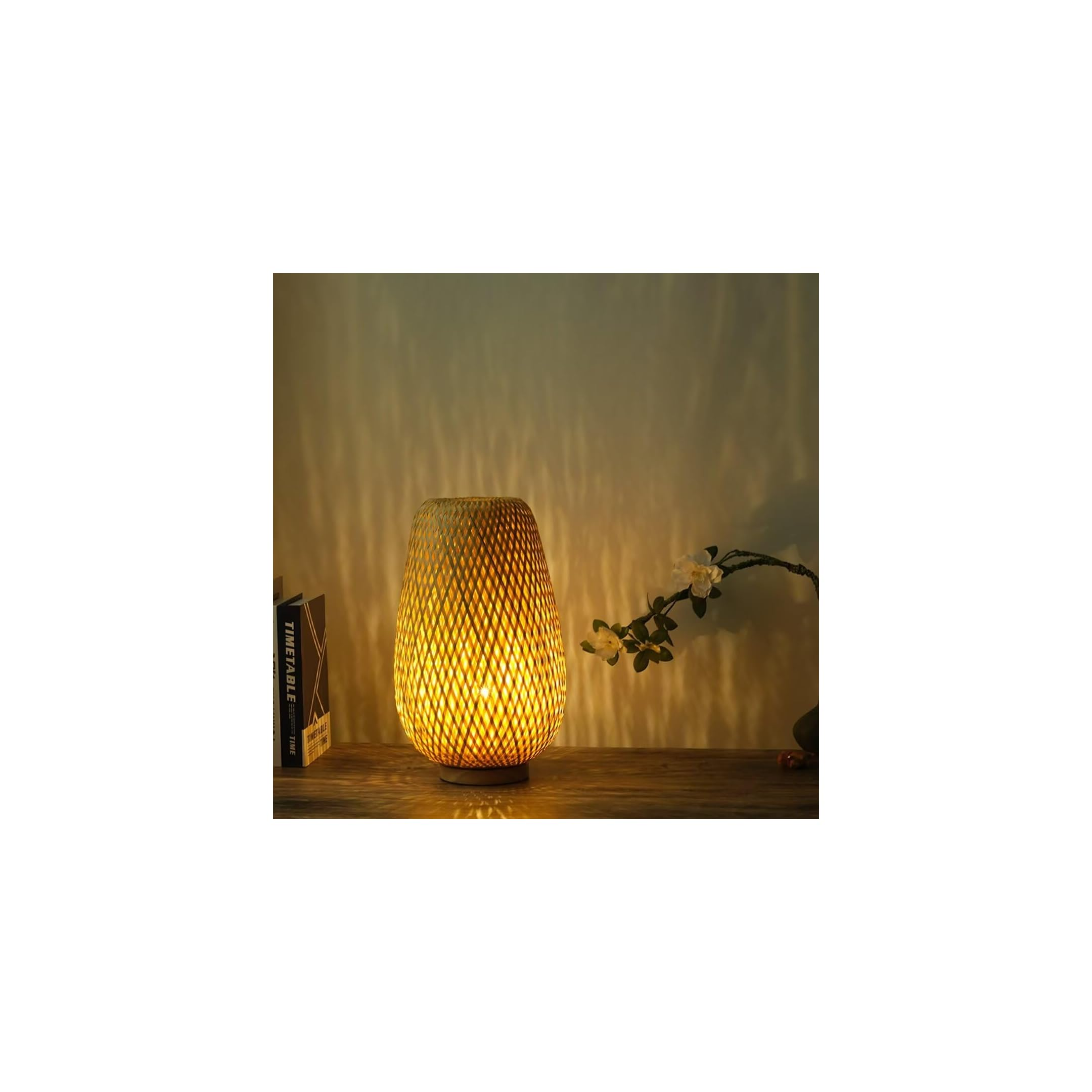Eine geflochtene Bambuslampe leuchtet auf einem Holz Tisch, im Hintergrund weiße Blüten und ein Buch.