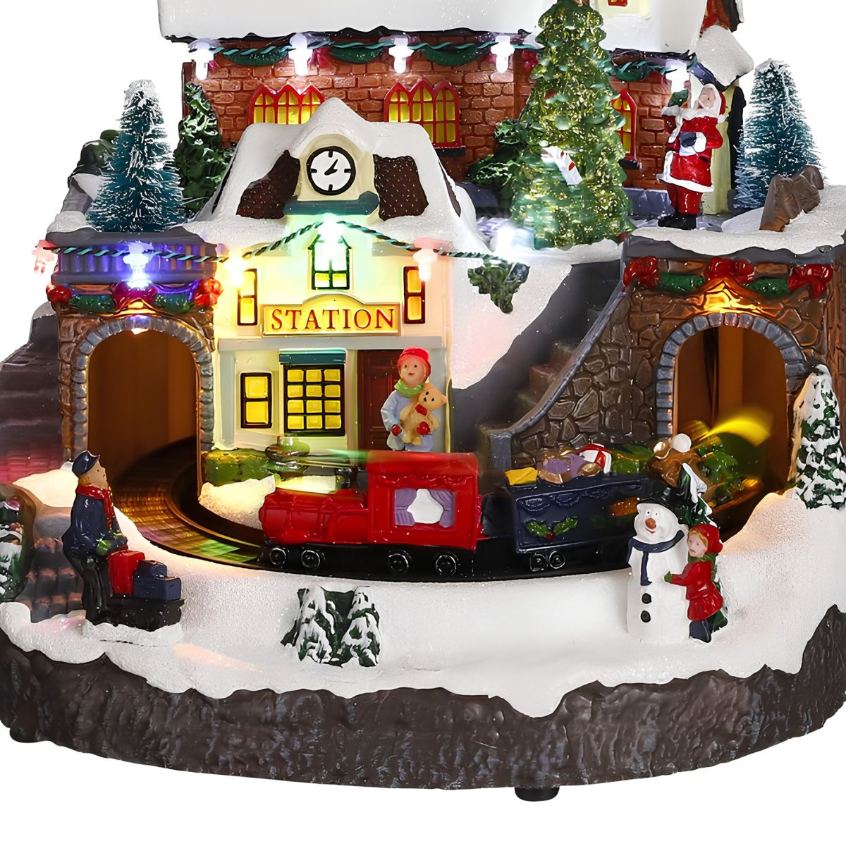 Verlicht kerstdorp met station, rode speelgoedtrein, besneeuwde figuren en bomen op een besneeuwde basis.
