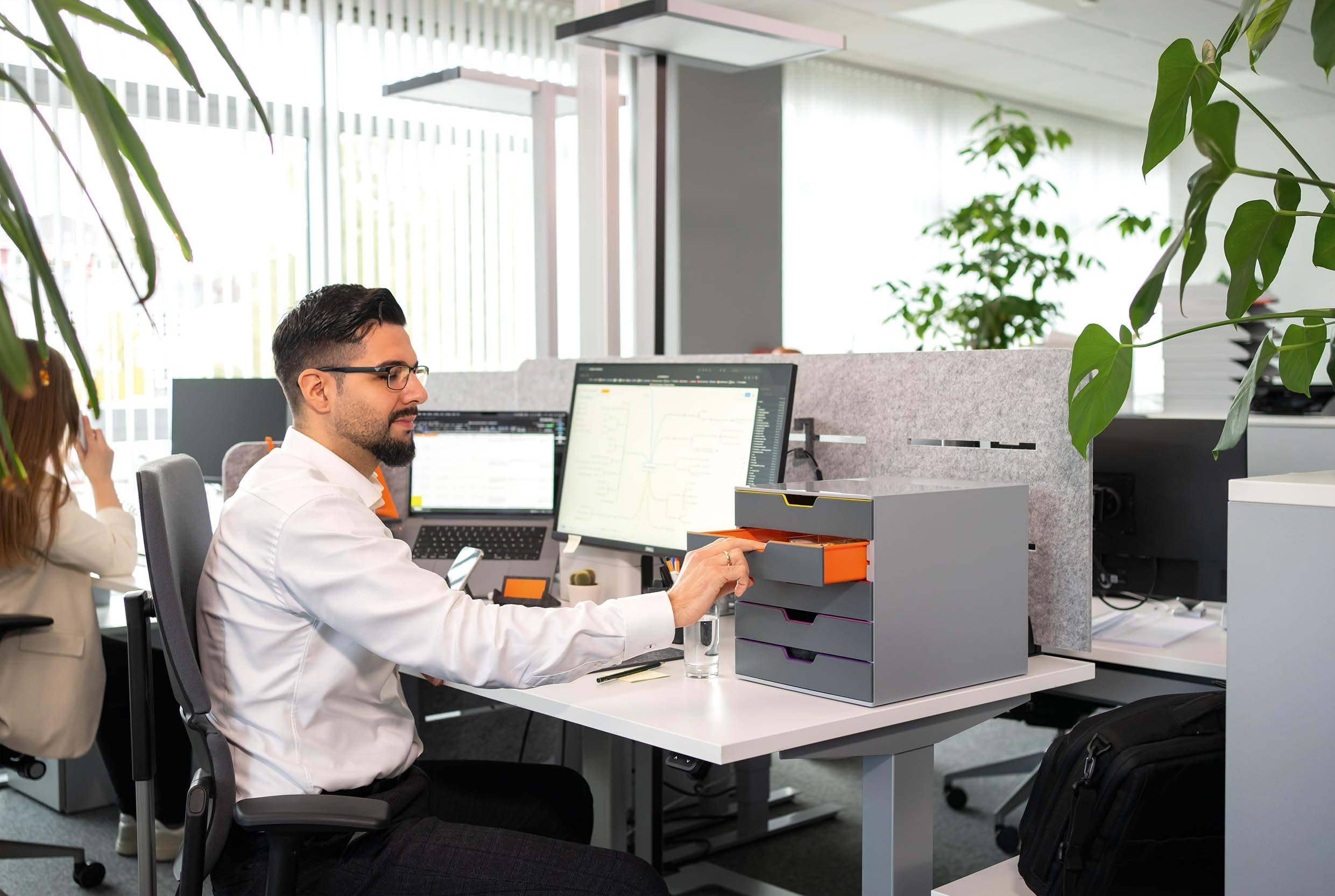 Ein Mann im weißen Hemd sitzt am Schreibtisch in einem modernen Büro und öffnet eine graue Schublade.