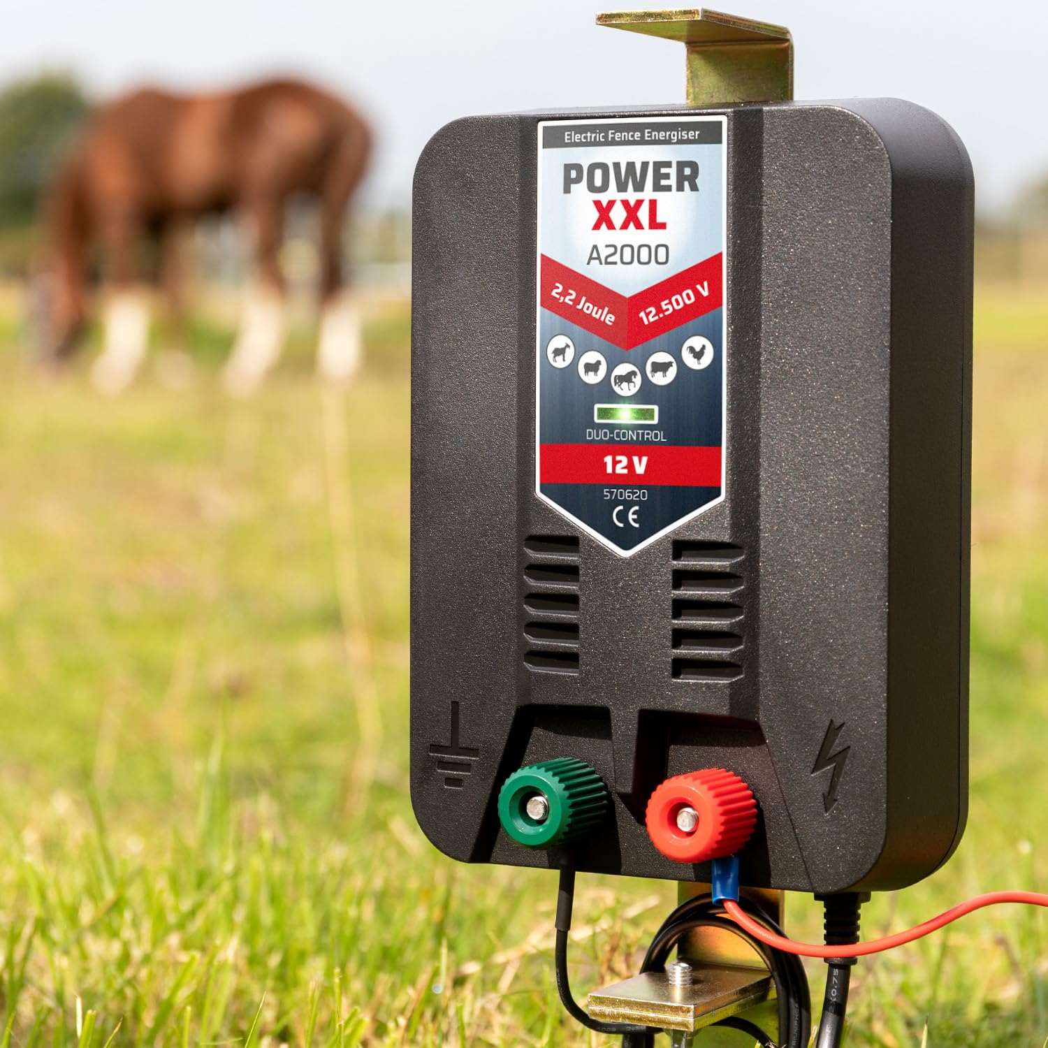 Un électrificateur de clôture noir avec des bornes rouges et vertes dans l'herbe, avec un cheval à l'arrière-plan.