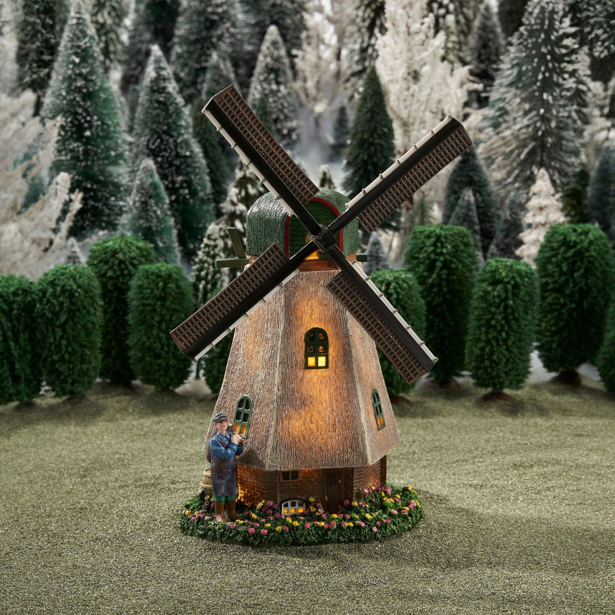 Een verlichte modelwindmolen met een figuur ernaast, tegen een achtergrond van miniatuurdennenbomen.