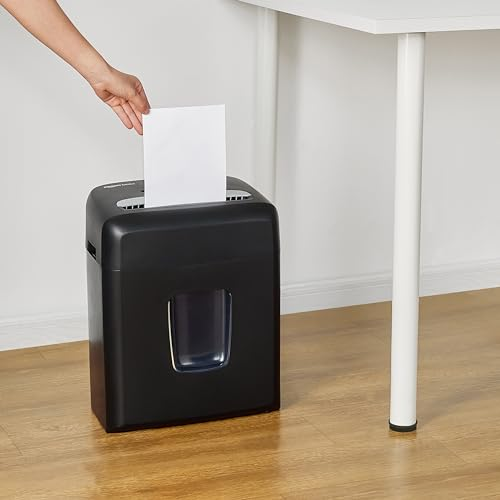 Eine Hand füttert Papier in einen schwarzen Aktenvernichter auf Holzboden neben einem Tisch.