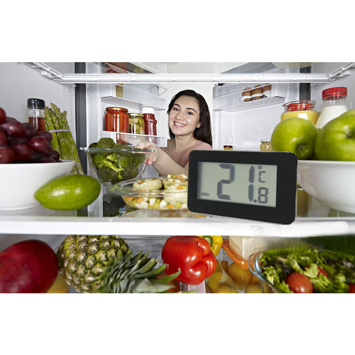 Ein Kühlschrank voller frischem Obst und Gemüse, mit einer Frau im Hintergrund und einem Digitalthermometer im Vordergrund.