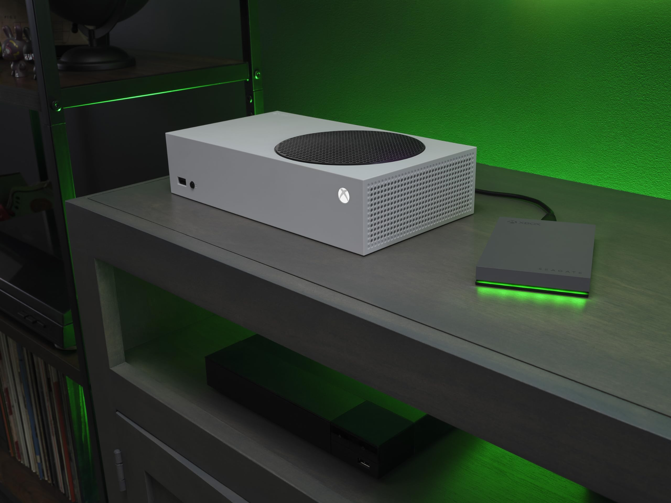 Eine weiße Xbox Series S und eine externe Seagate-Festplatte liegen auf einer dunkelgrauen Medienkonsole vor einer grünen Wand.