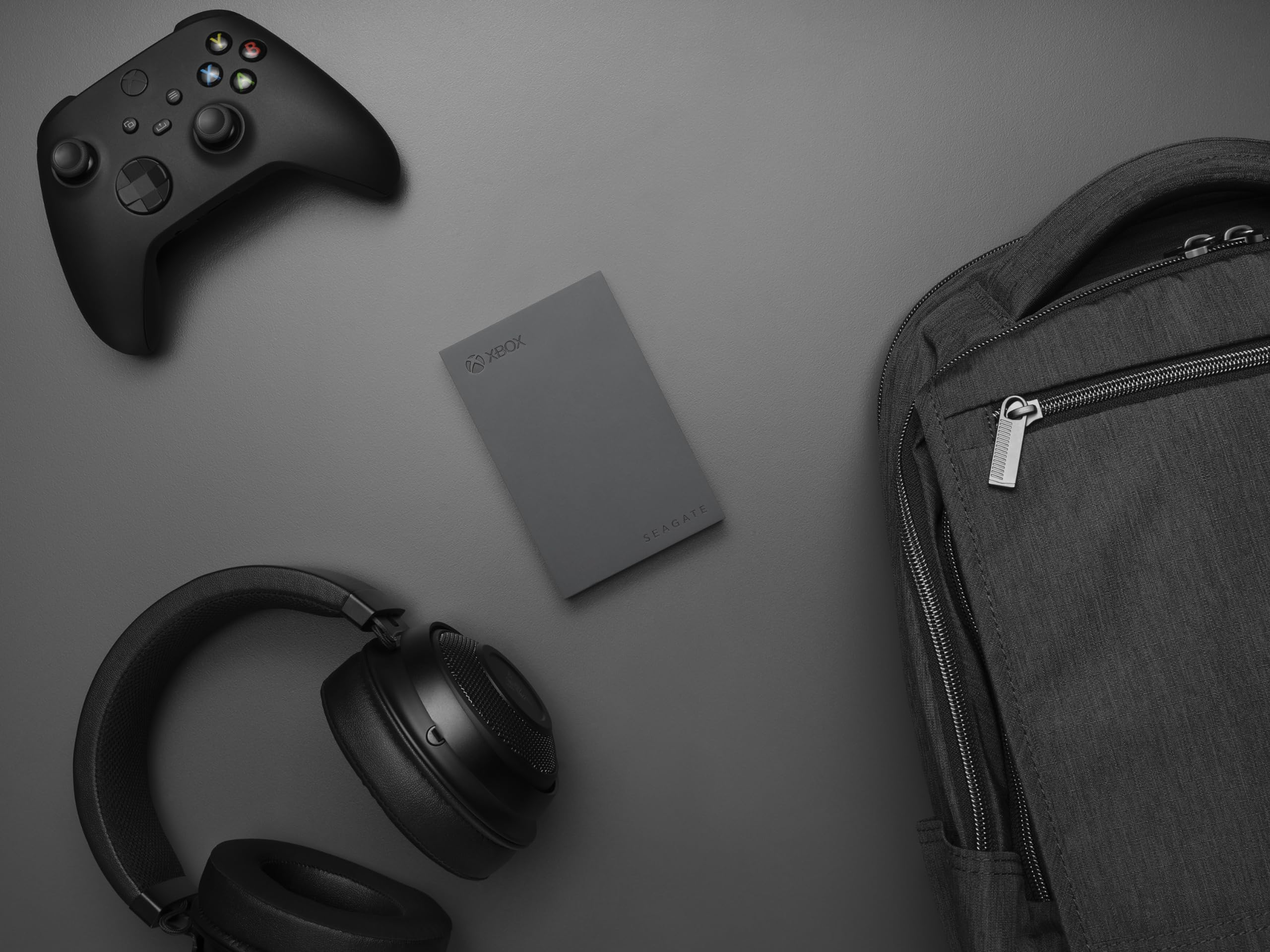 Ein schwarzer Xbox-Controller, Kopfhörer, eine Seagate-Festplatte und ein Rucksack auf einer grauen Oberfläche.